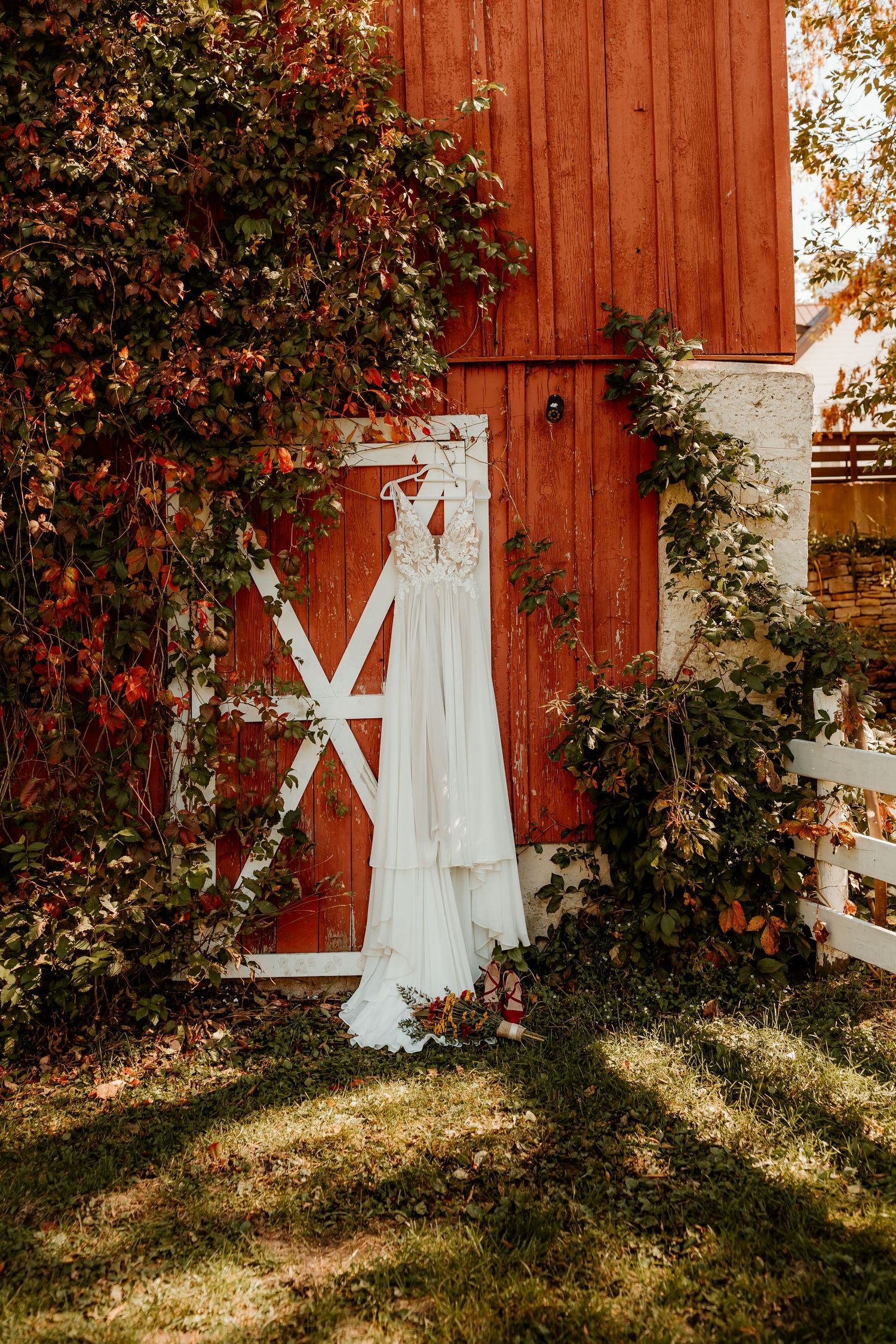 White lace wedding dress hanging on a rustic red barn door surrounded by autumn vines, with a small bouquet resting on the grass below.