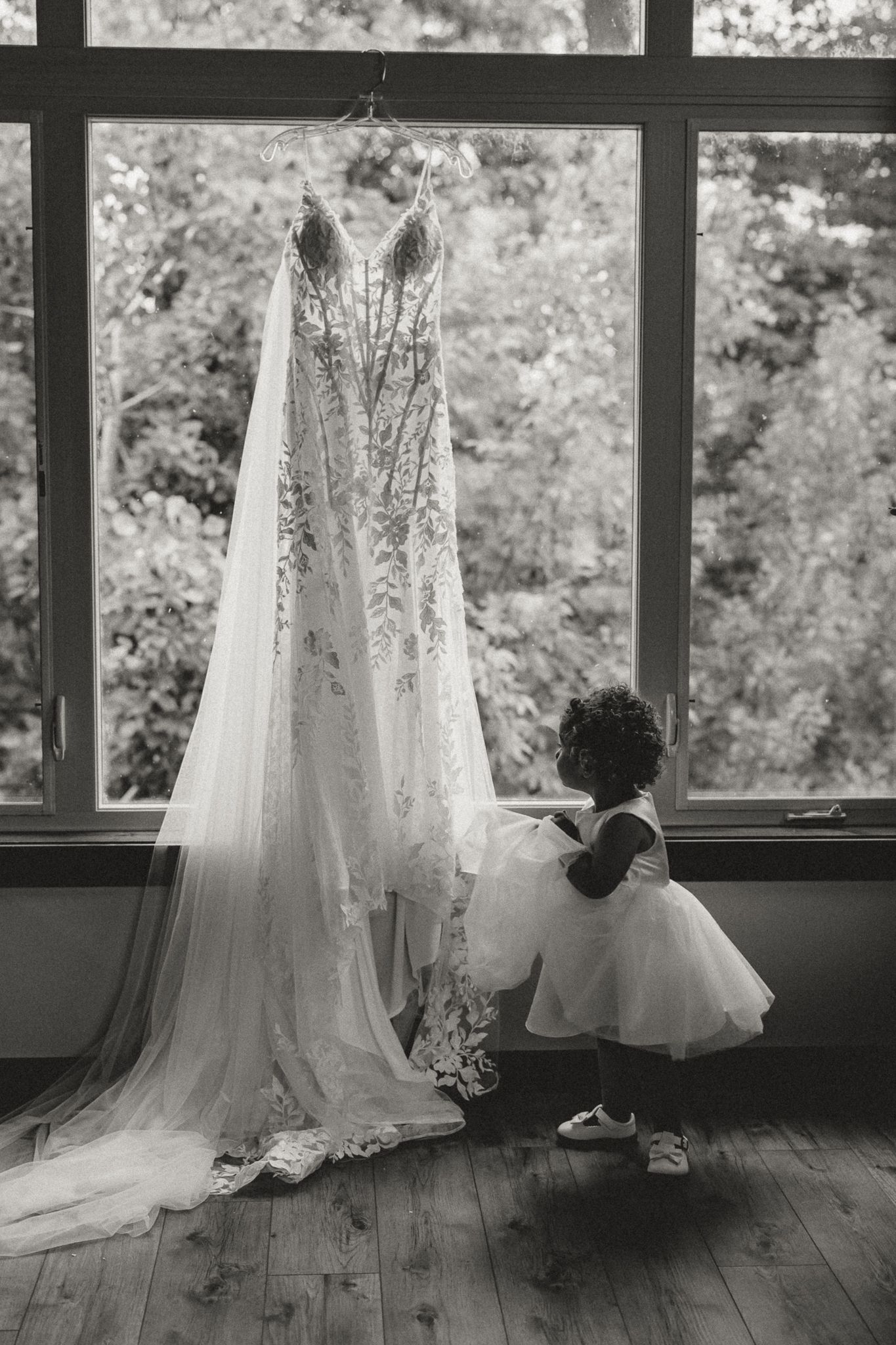 Black and white photo of a toddler in a tutu dress reaching toward a lace wedding gown with a long veil hanging by a window.