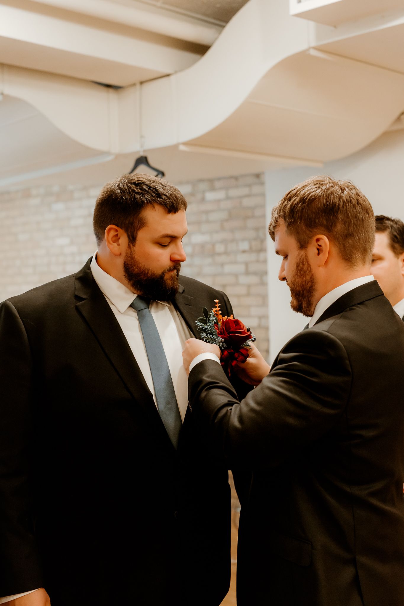 Groomsman in a dark suit pins a red floral boutonniere onto a bearded man's lapel before a wedding in a bright room with exposed brick.