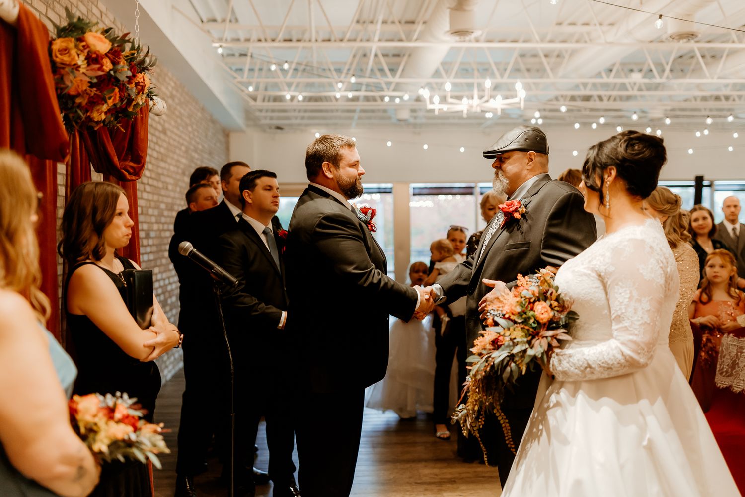 Groom in a black suit shakes hands with an older man as a bride in a white gown holds a bouquet during a wedding ceremony.