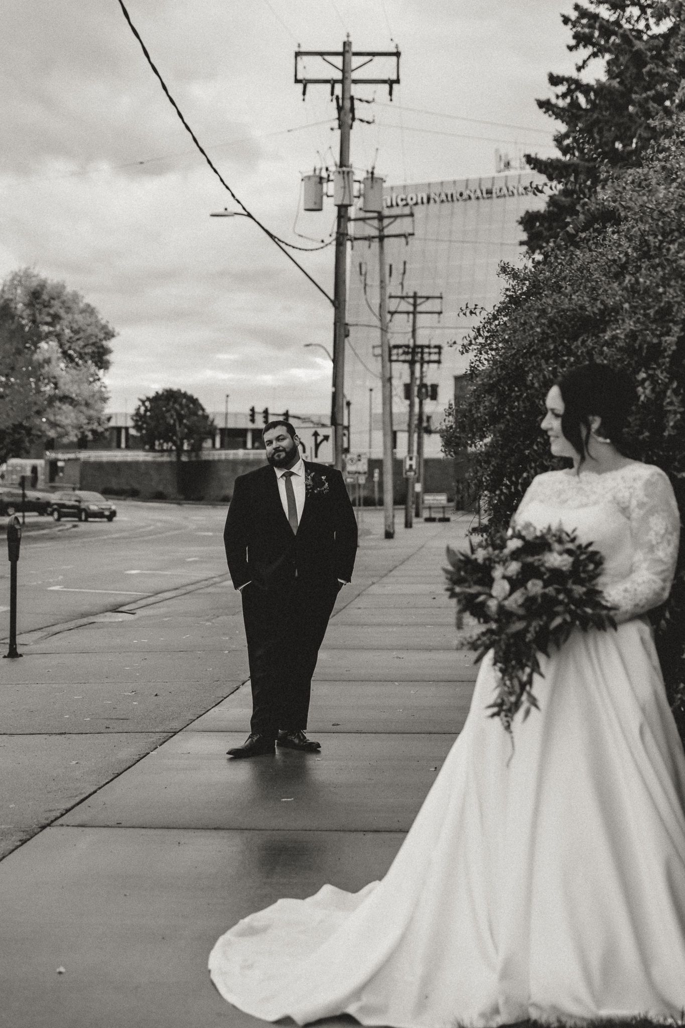 Black and white photo of a bride in a lace gown holding a bouquet as the groom in a suit stands on a city sidewalk beside utility poles.
