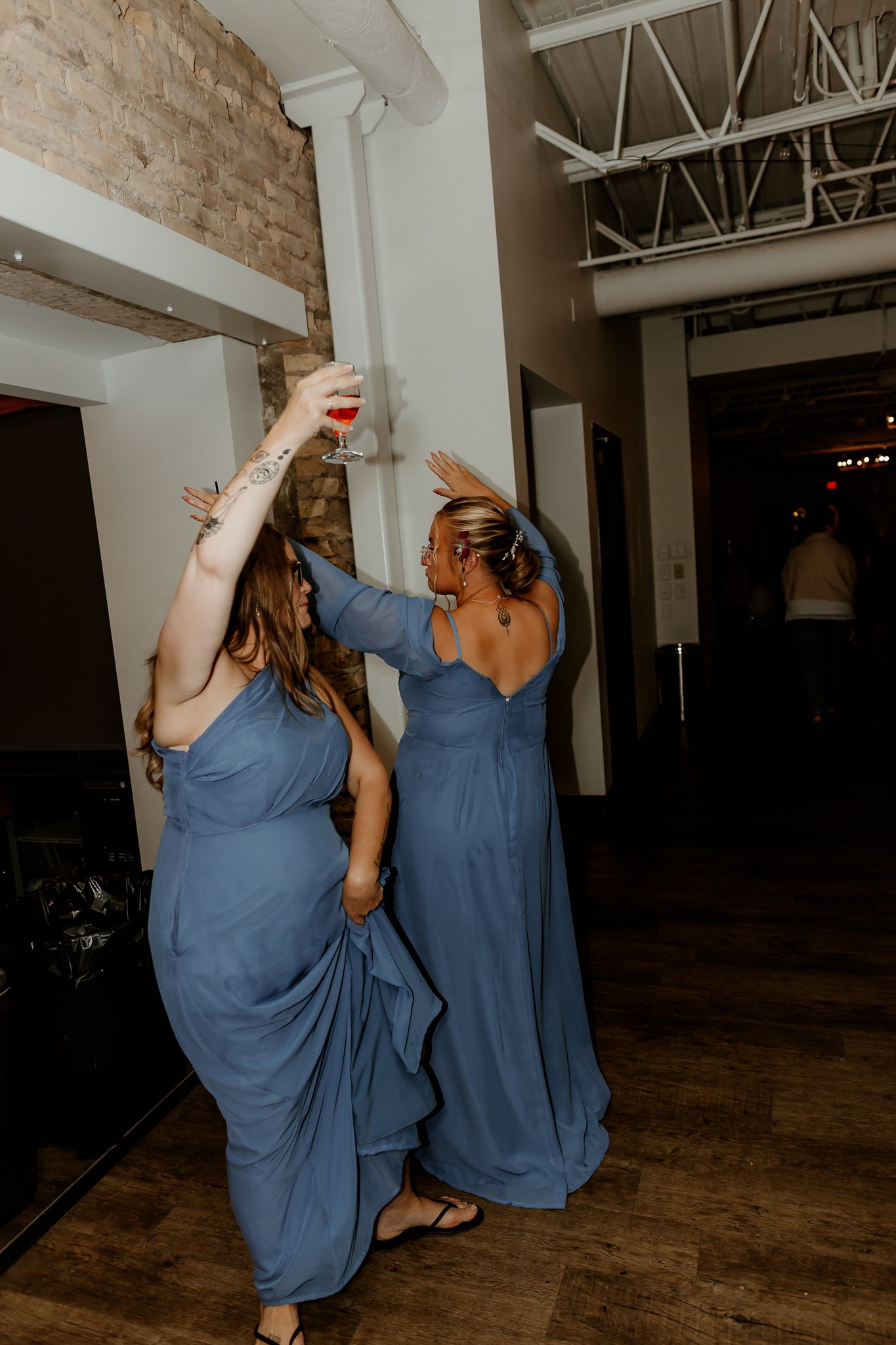 Two women in matching dusty blue dresses dance with arms raised, one holding a drink, in a brick-walled venue with exposed ceiling beams.