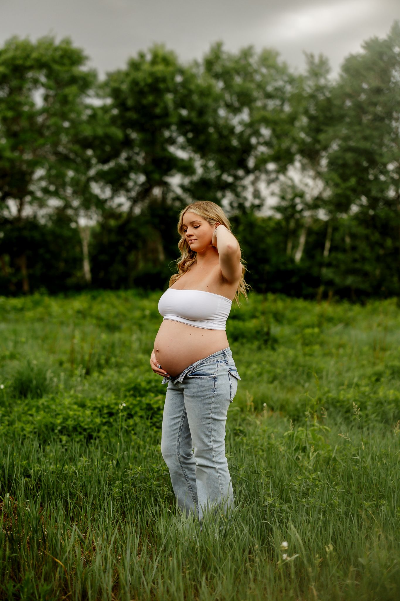 Pregnant woman in white bandeau top and low-rise jeans standing in lush green field at dusk.