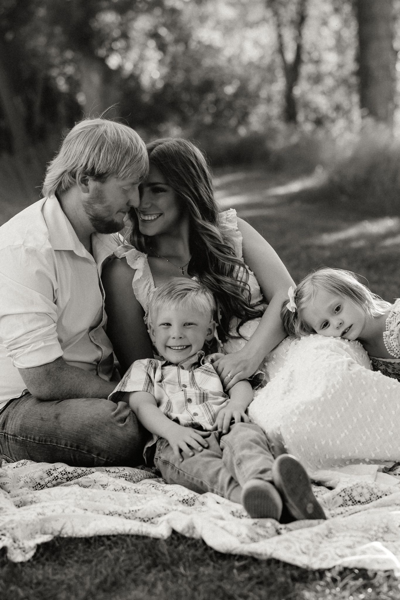 Black and white photo of a loving family of four sitting together on a blanket under trees.