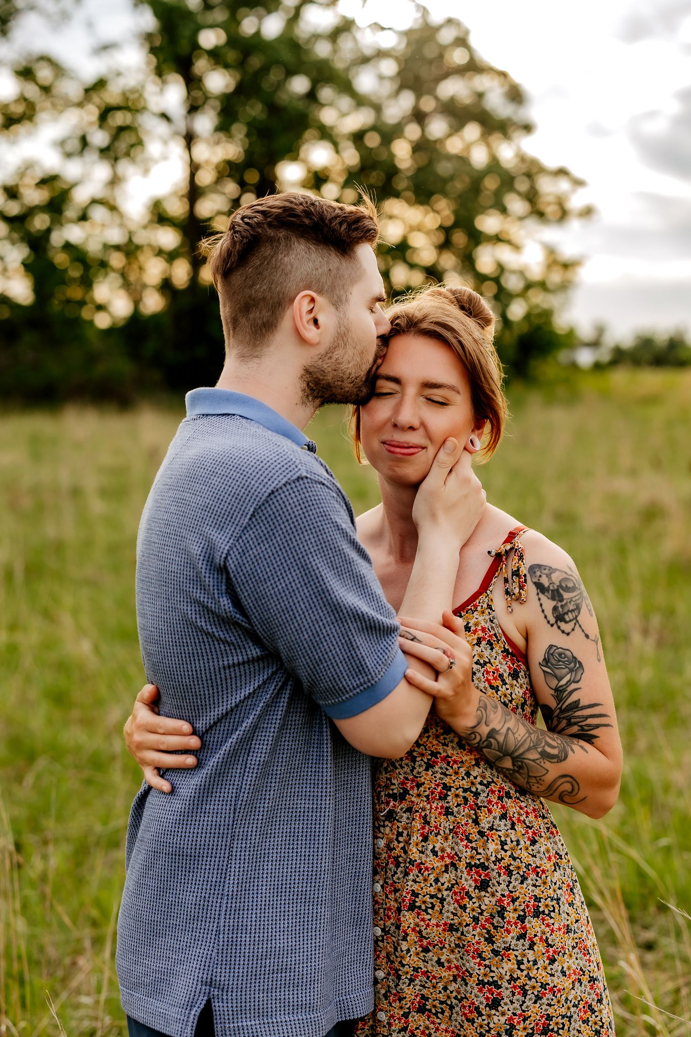 A man kisses a woman on the cheek as she smiles with eyes closed in a sunlit green field.