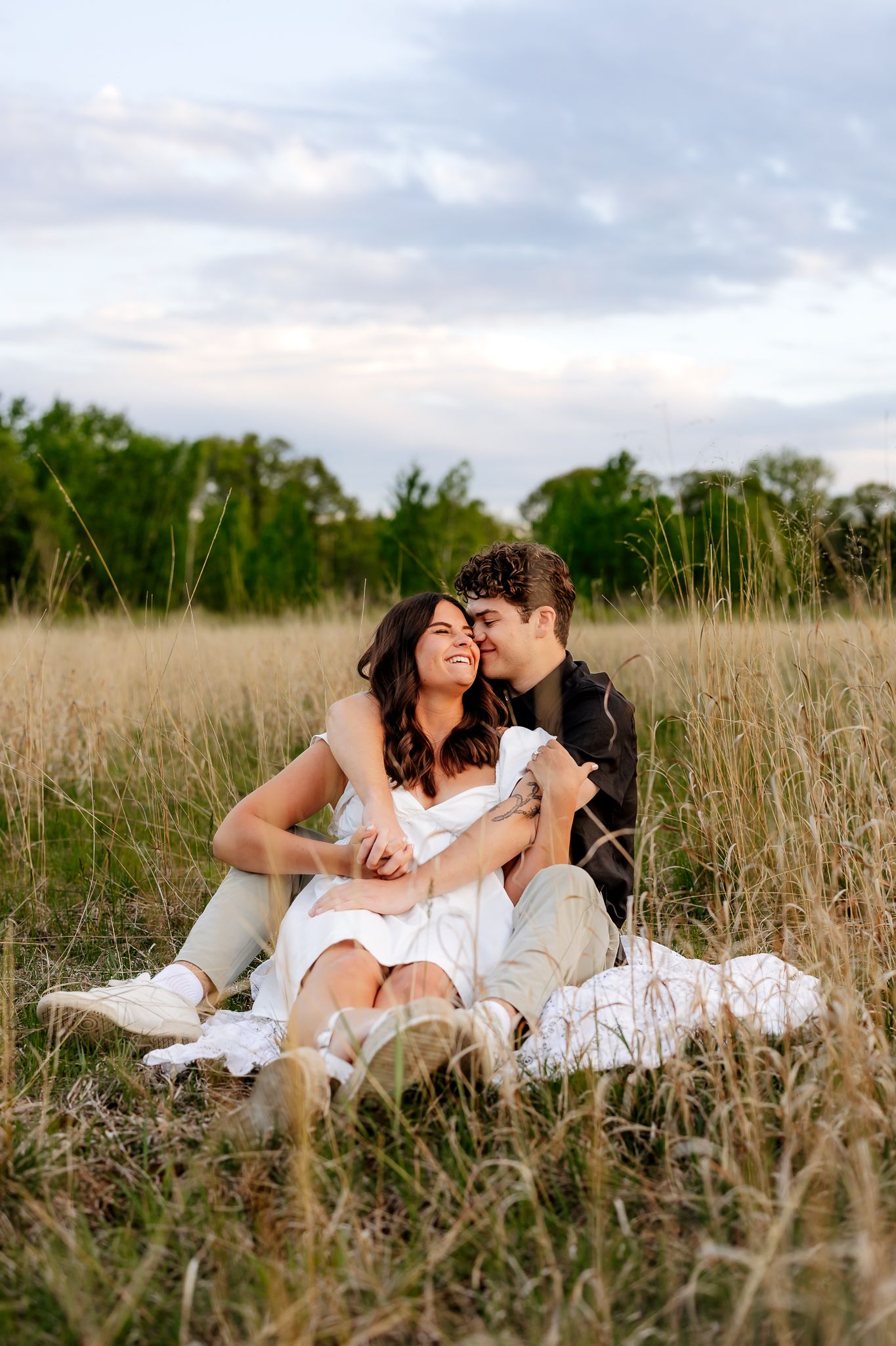 Romantic couple sitting together in tall grass field at dusk, sharing a tender moment.