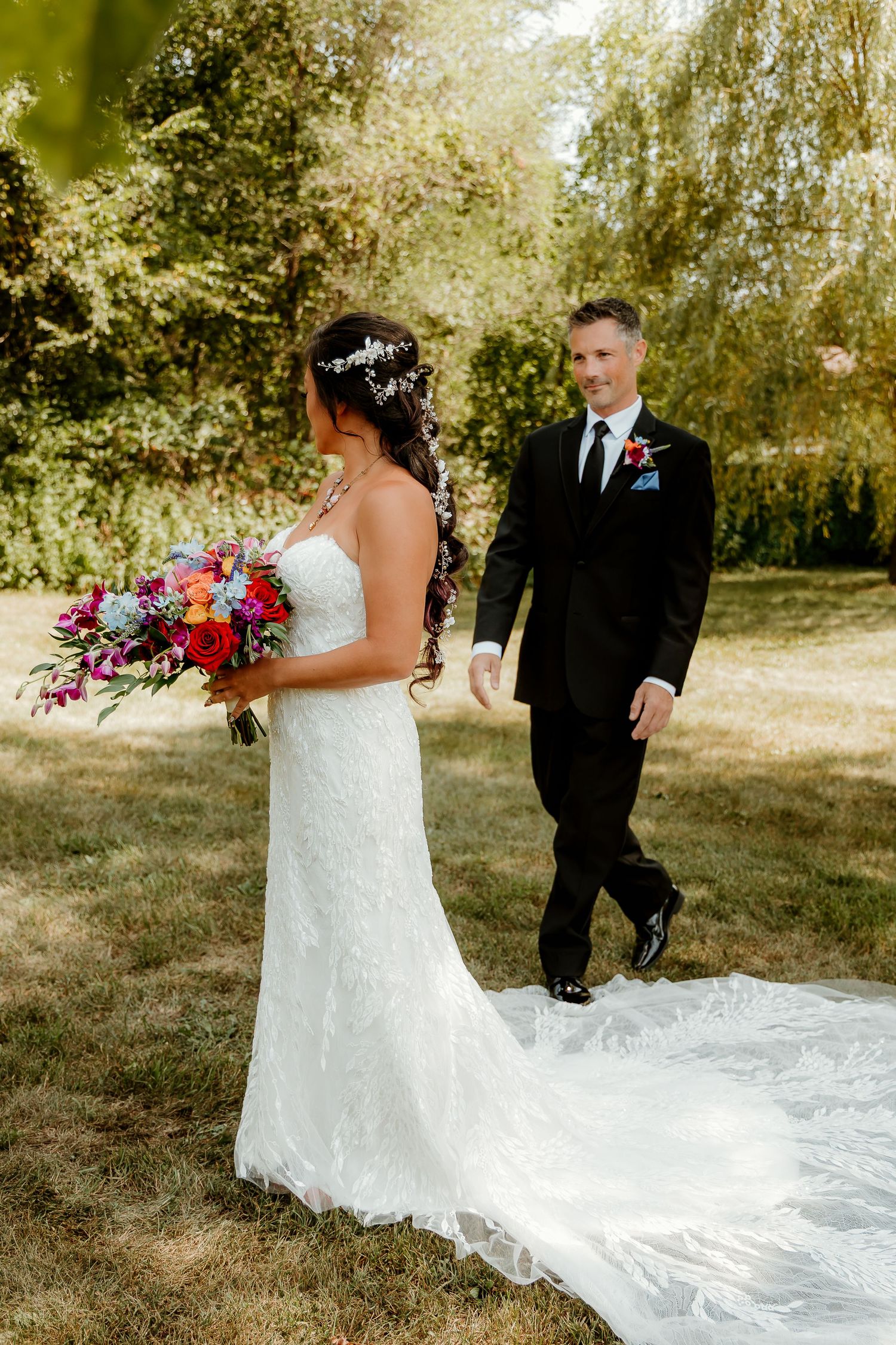 Bride in white lace gown holding colorful bouquet faces groom in black suit walking toward her in sunny garden.