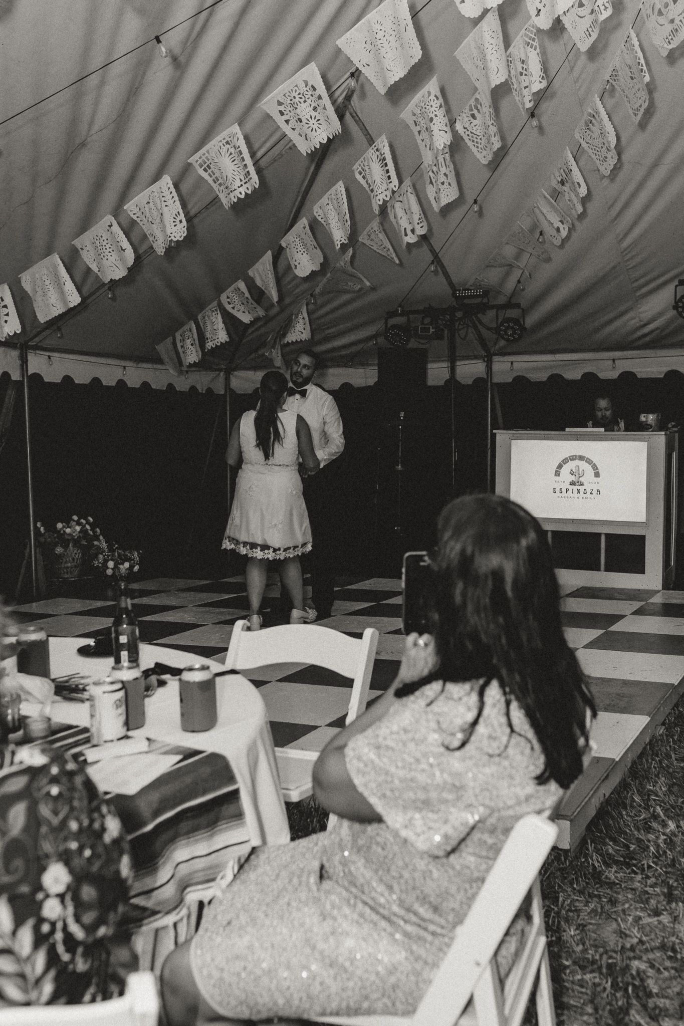 Black and white photo of a wedding reception under a tent with papel picado decorations, checkered dance floor, and guests seated at tables.