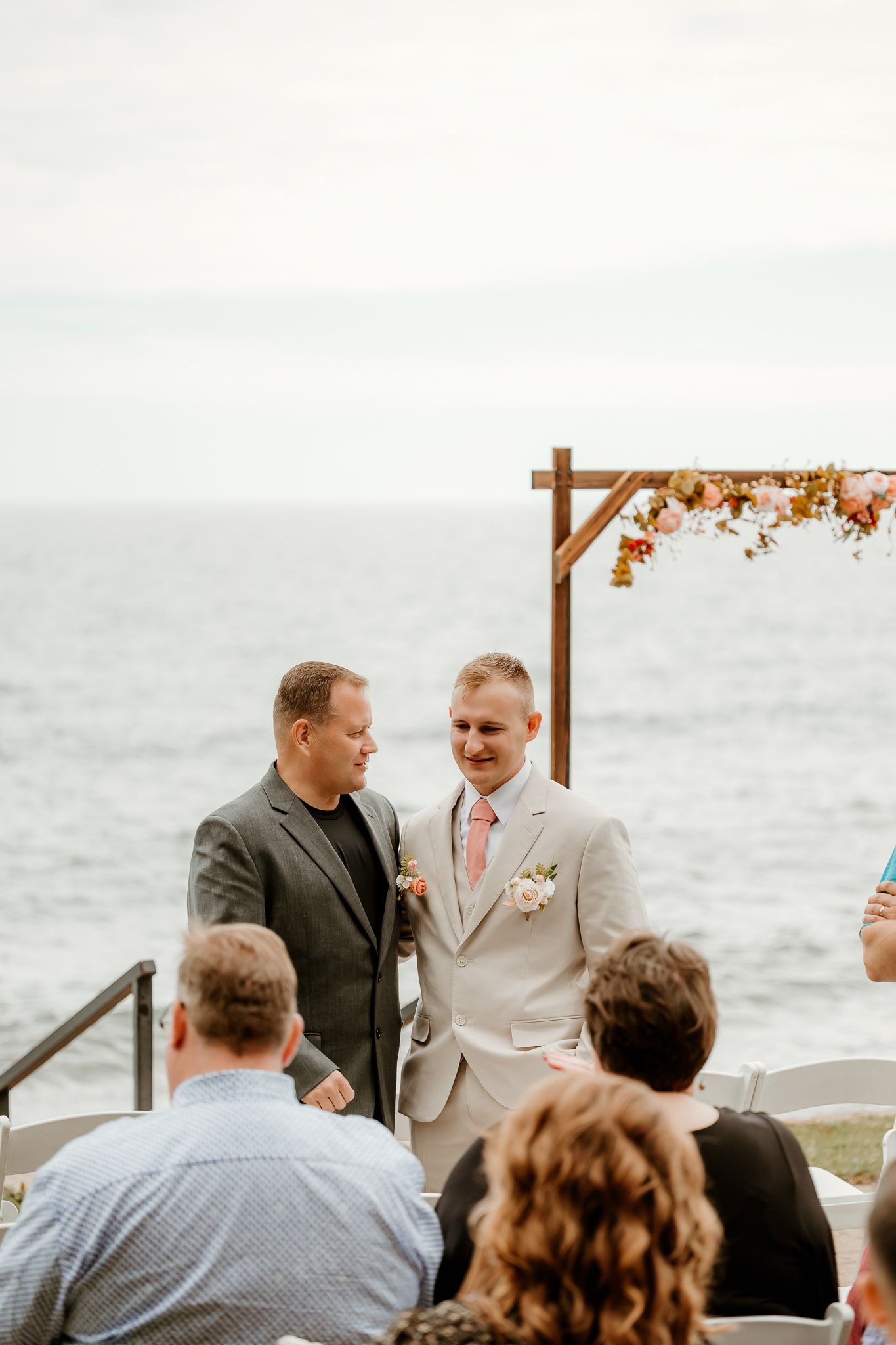 Two grooms share a joyful moment at an outdoor oceanside wedding ceremony beneath a floral arch.