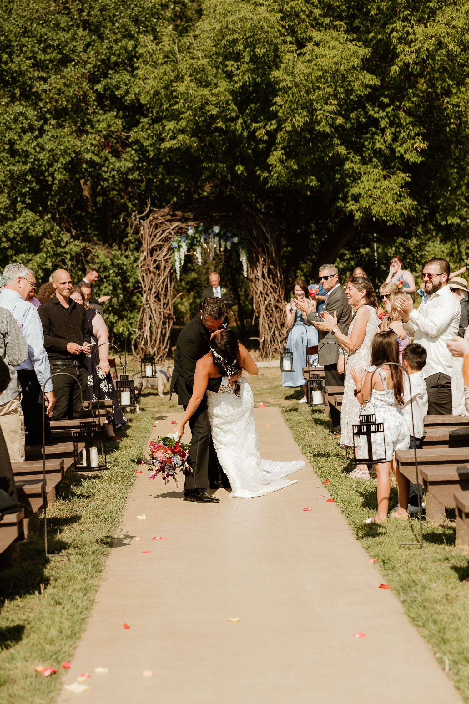 Bride and groom share a kiss at outdoor wedding ceremony aisle lined with lanterns and rose petals under lush green trees.
