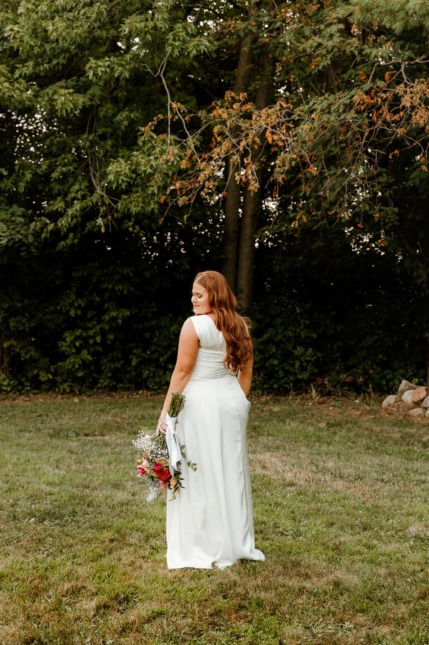 Bride in white gown holding colorful bouquet stands on green lawn before lush trees, hair flowing down her back.
