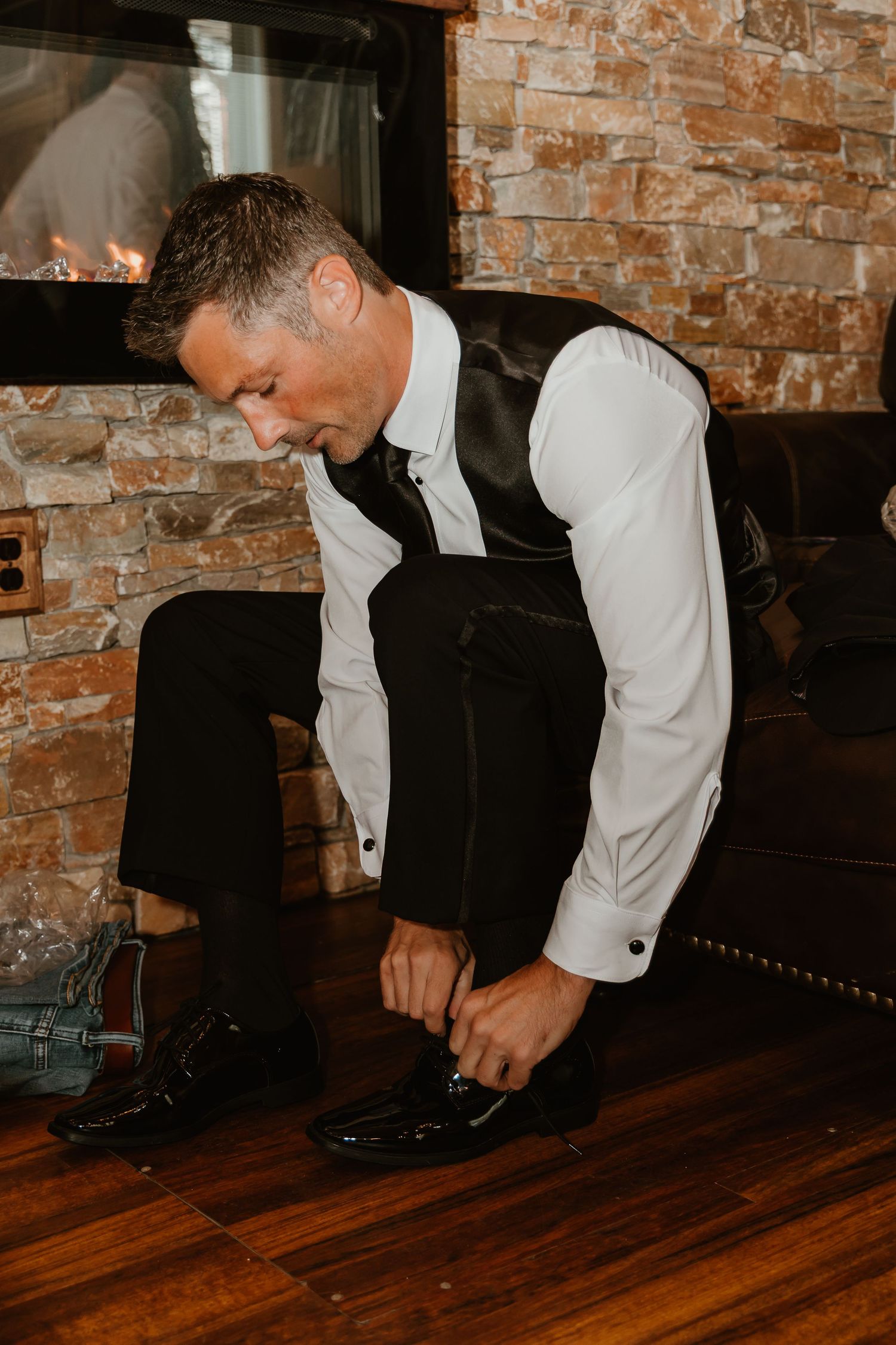 A groom in a black vest and white dress shirt ties his shiny black shoes near a brick fireplace on hardwood floors.