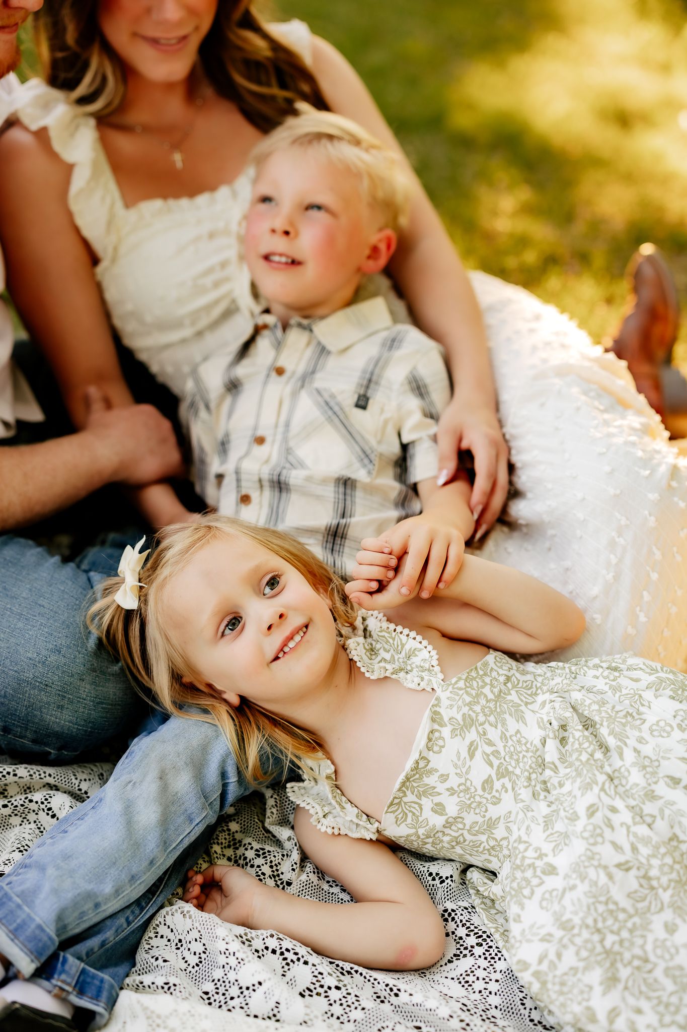 Two young siblings smile during a golden hour outdoor family photo session on a lace blanket in green grass.