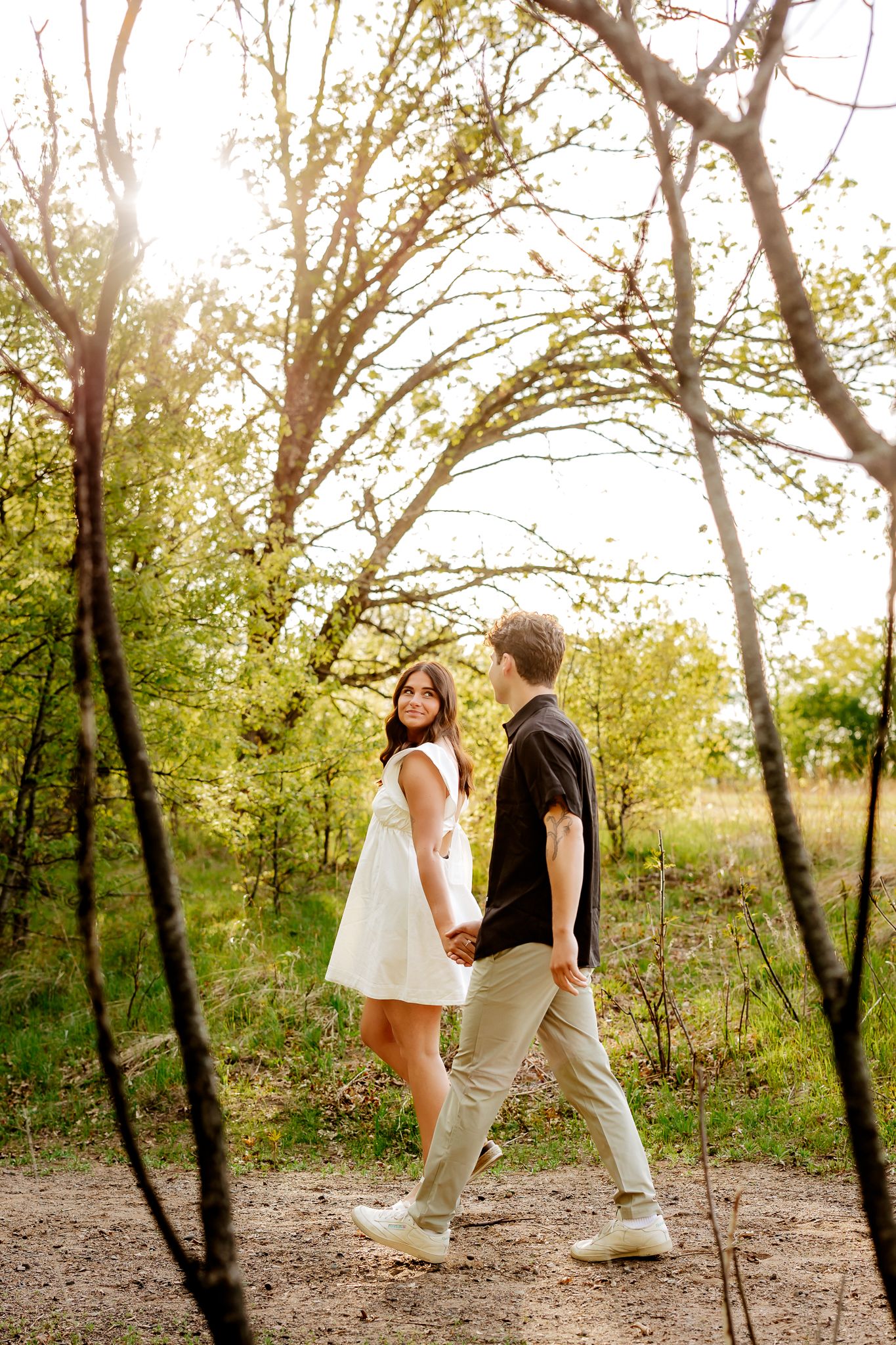 Couple holds hands walking through sunlit forest path, woman in white dress looks back smiling.