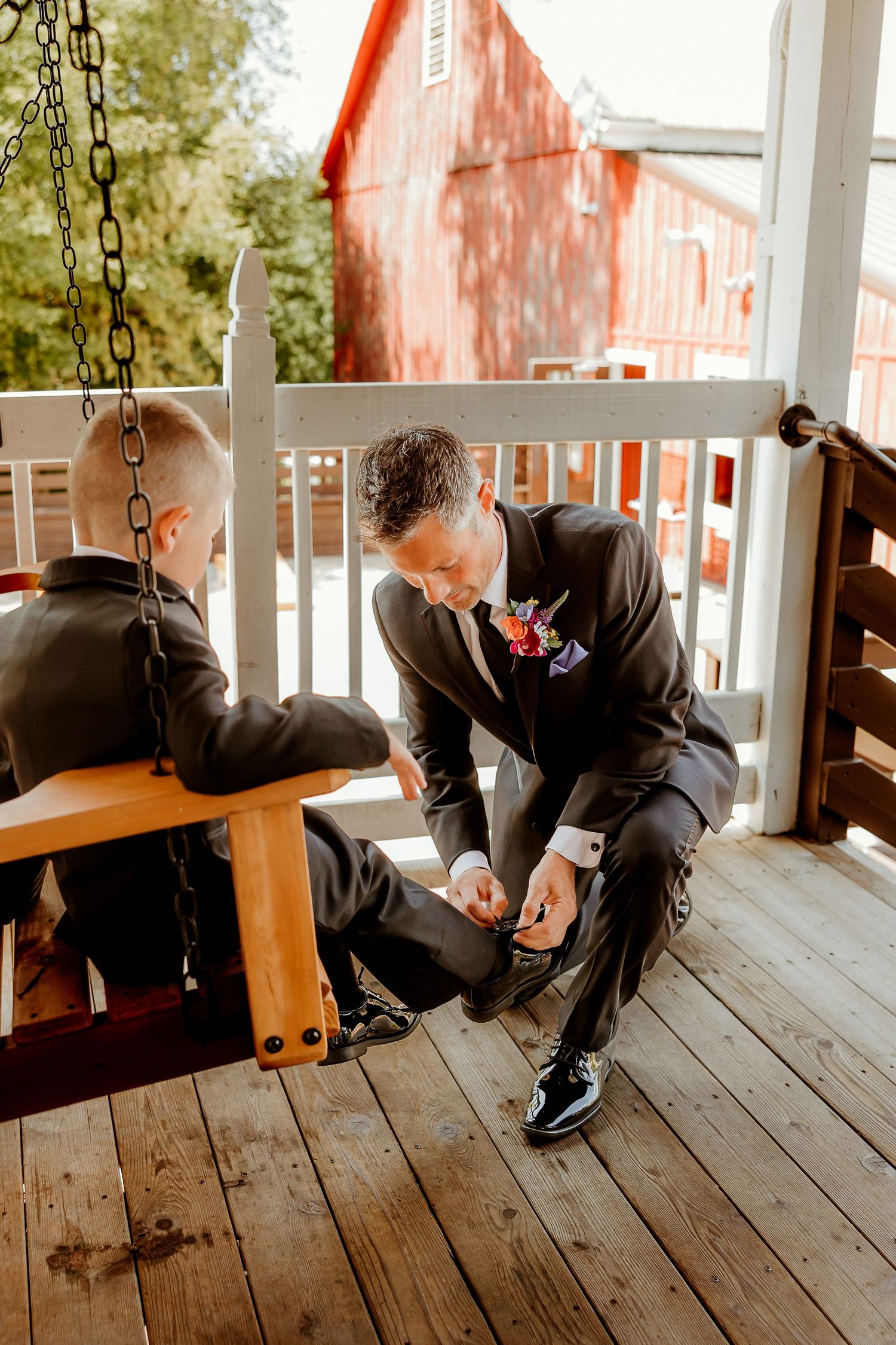 Groom in dark suit helps young boy with shoes on wooden porch swing, red barn visible in background.