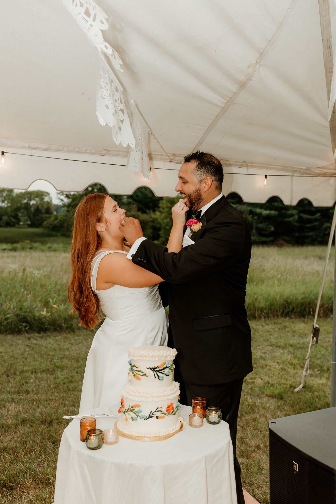 Bride and groom feeding each other wedding cake under a white tent at an outdoor reception.