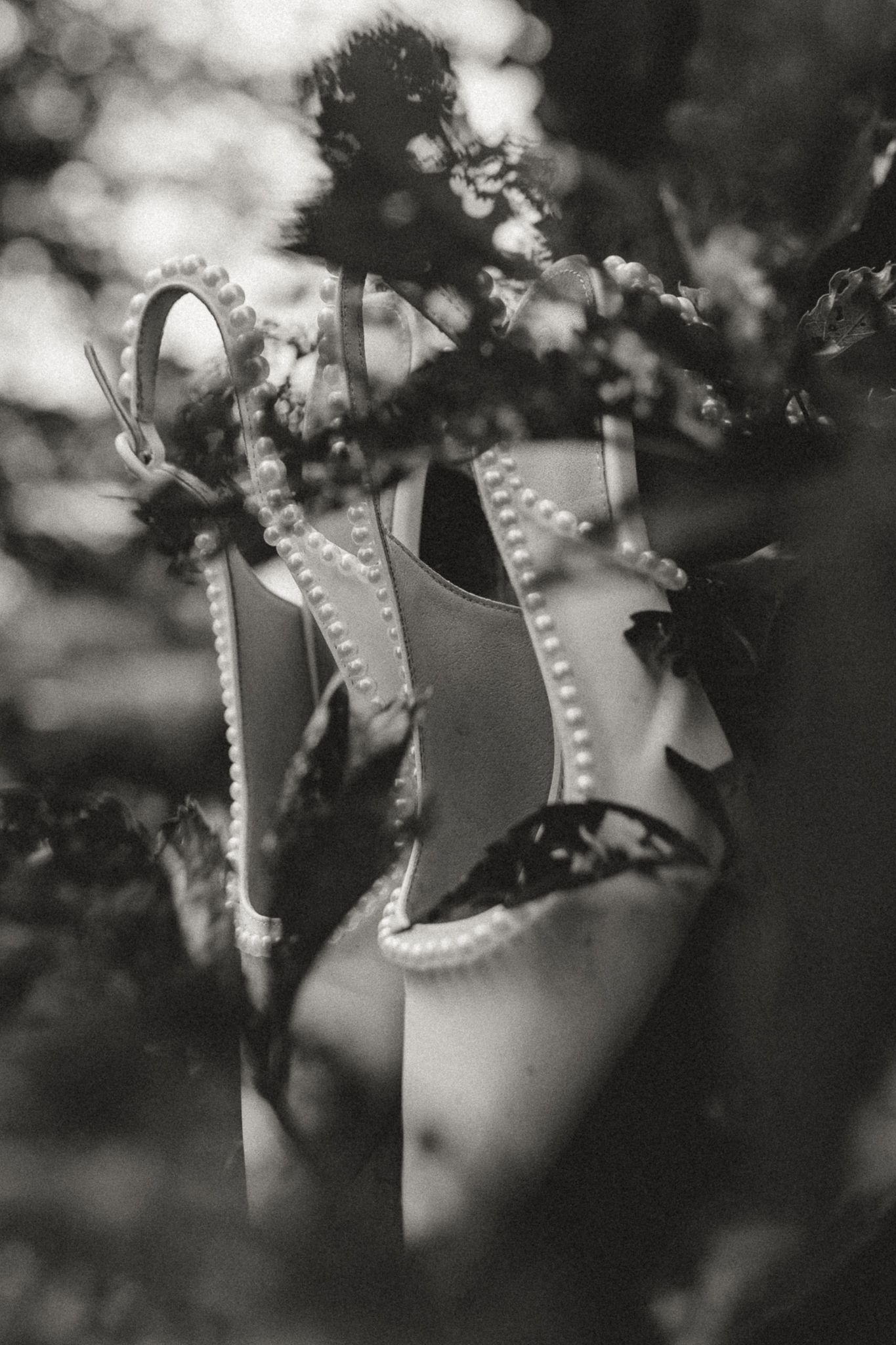 Black and white photo of elegant pearl-embellished bridal heels nestled among lush garden foliage.