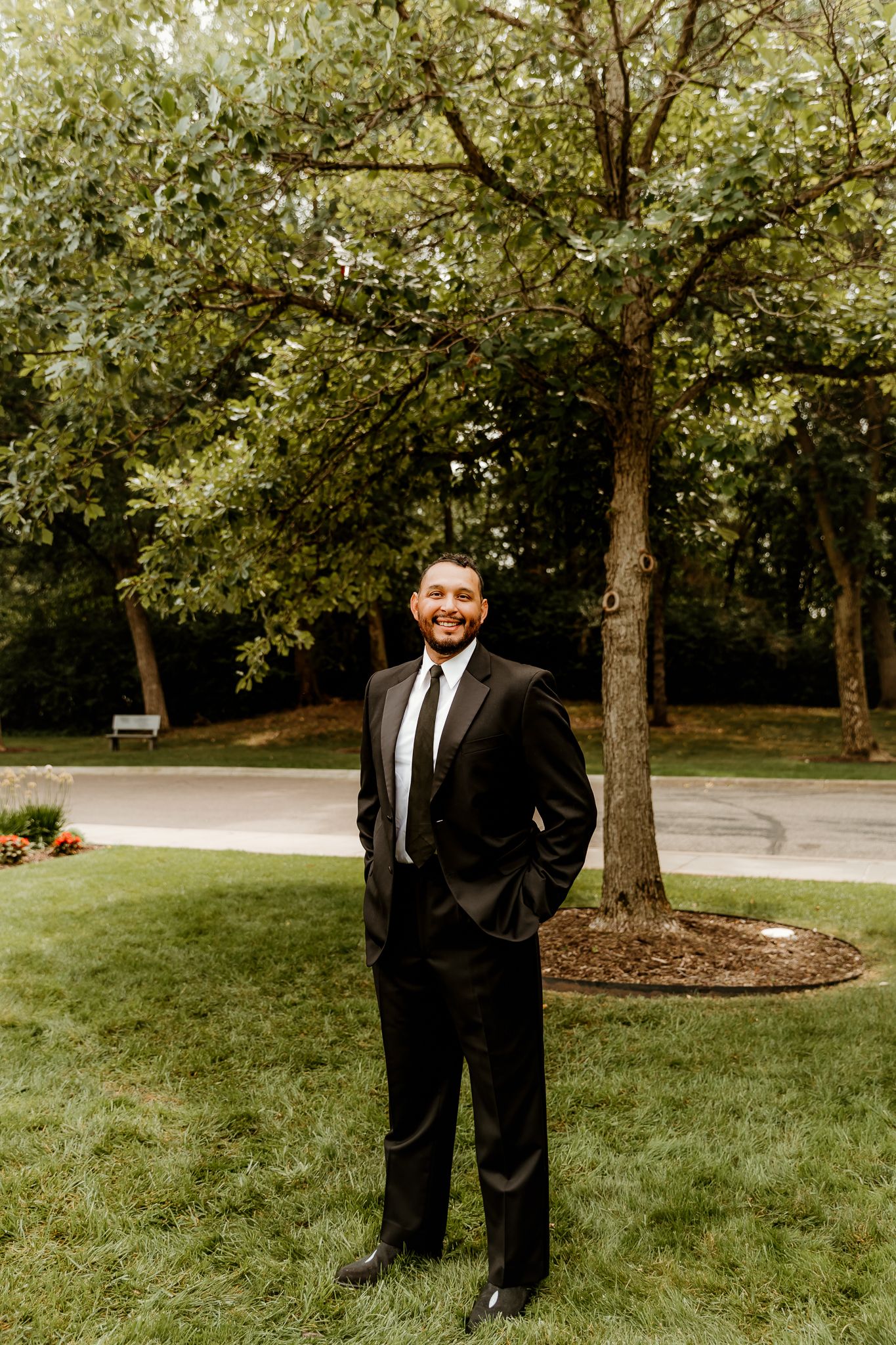 A smiling man in a black suit and tie stands on green grass in front of a large tree outdoors.