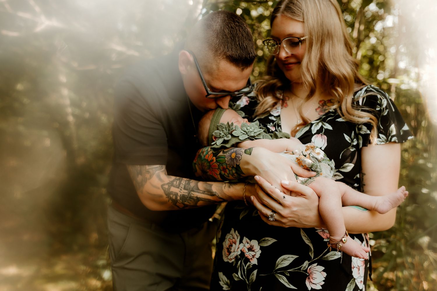 A tattooed father kisses his newborn baby while mother holds the infant during an outdoor family photo session.