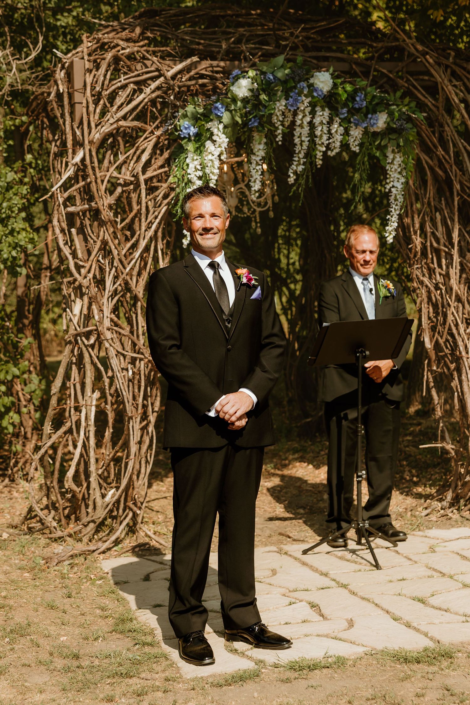 Groom in black suit smiling at altar under rustic branch arch with blue flowers, officiant standing behind at outdoor wedding.