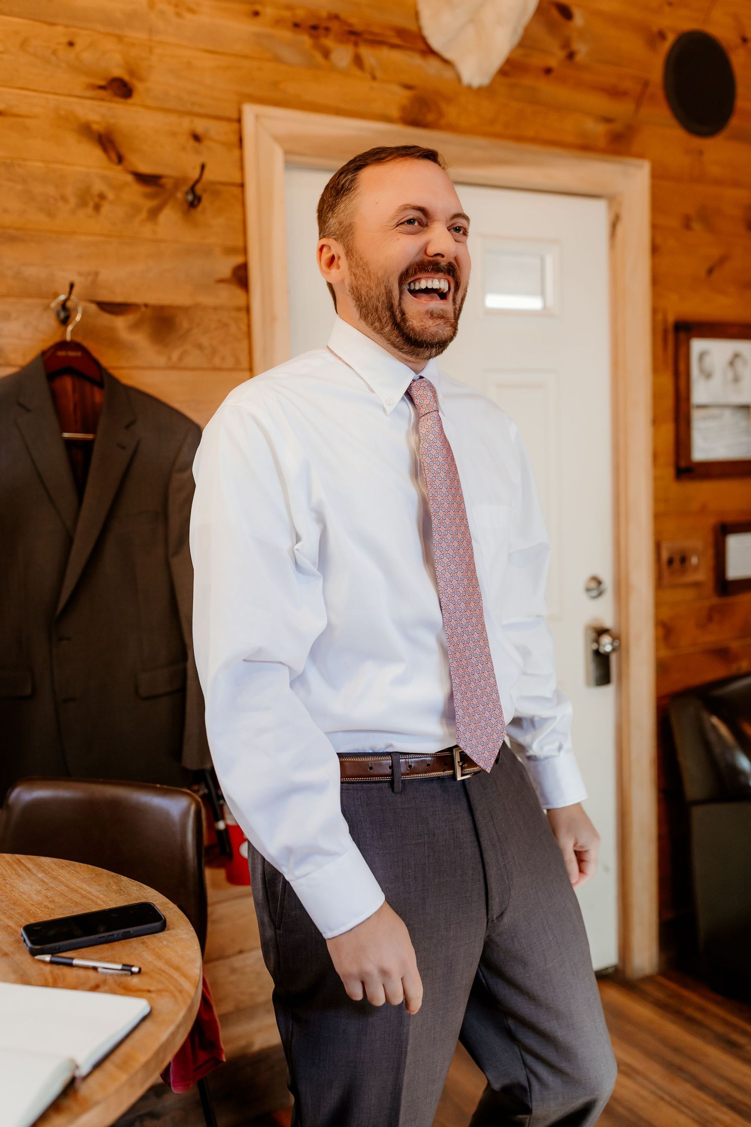 Smiling bearded man in a white dress shirt and pink patterned tie laughs in a rustic wood cabin, with a suit jacket hanging nearby.