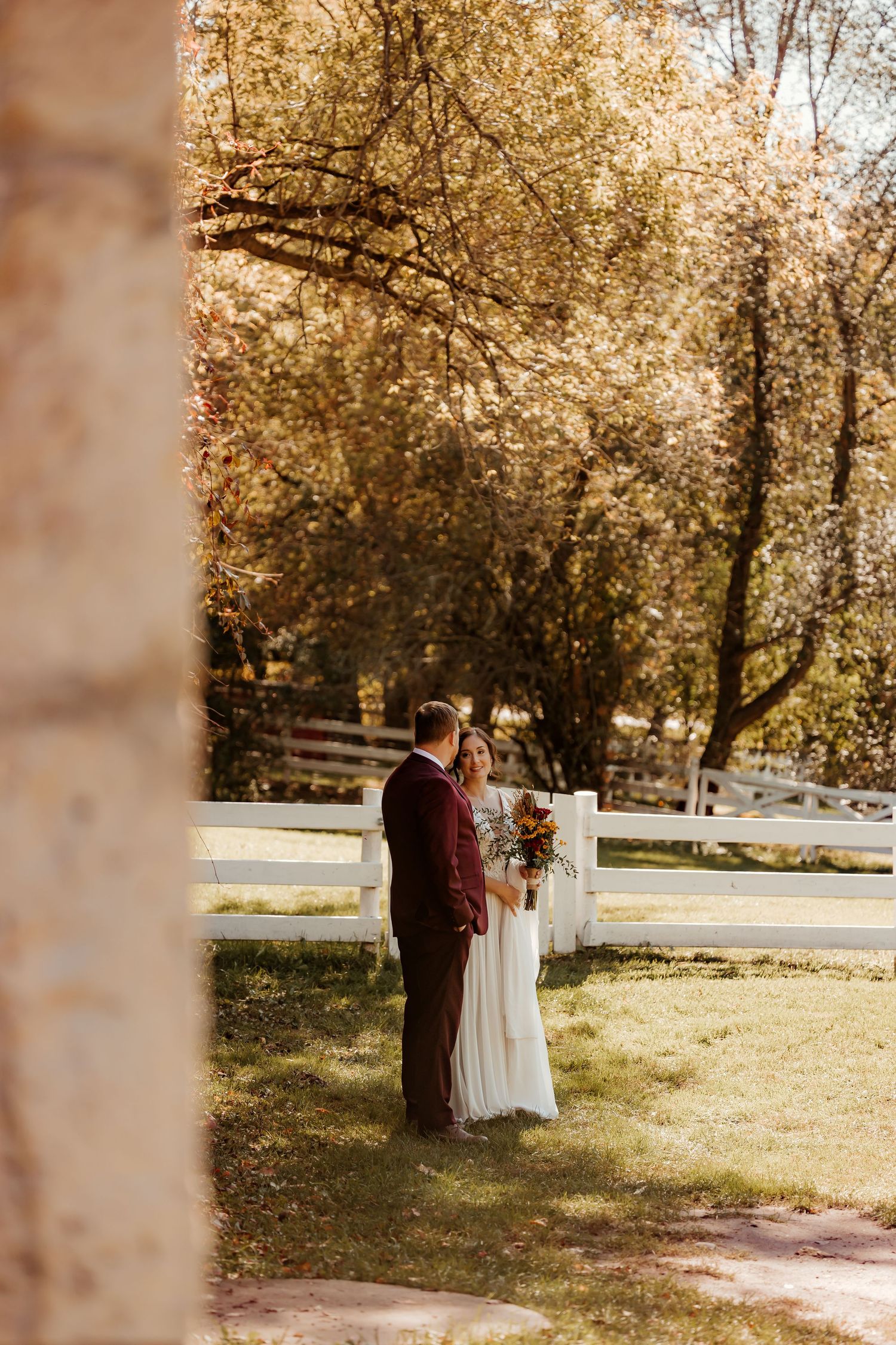 Bride in a white gown holding a bouquet stands beside a groom in a burgundy suit near a white fence, framed by autumn trees.