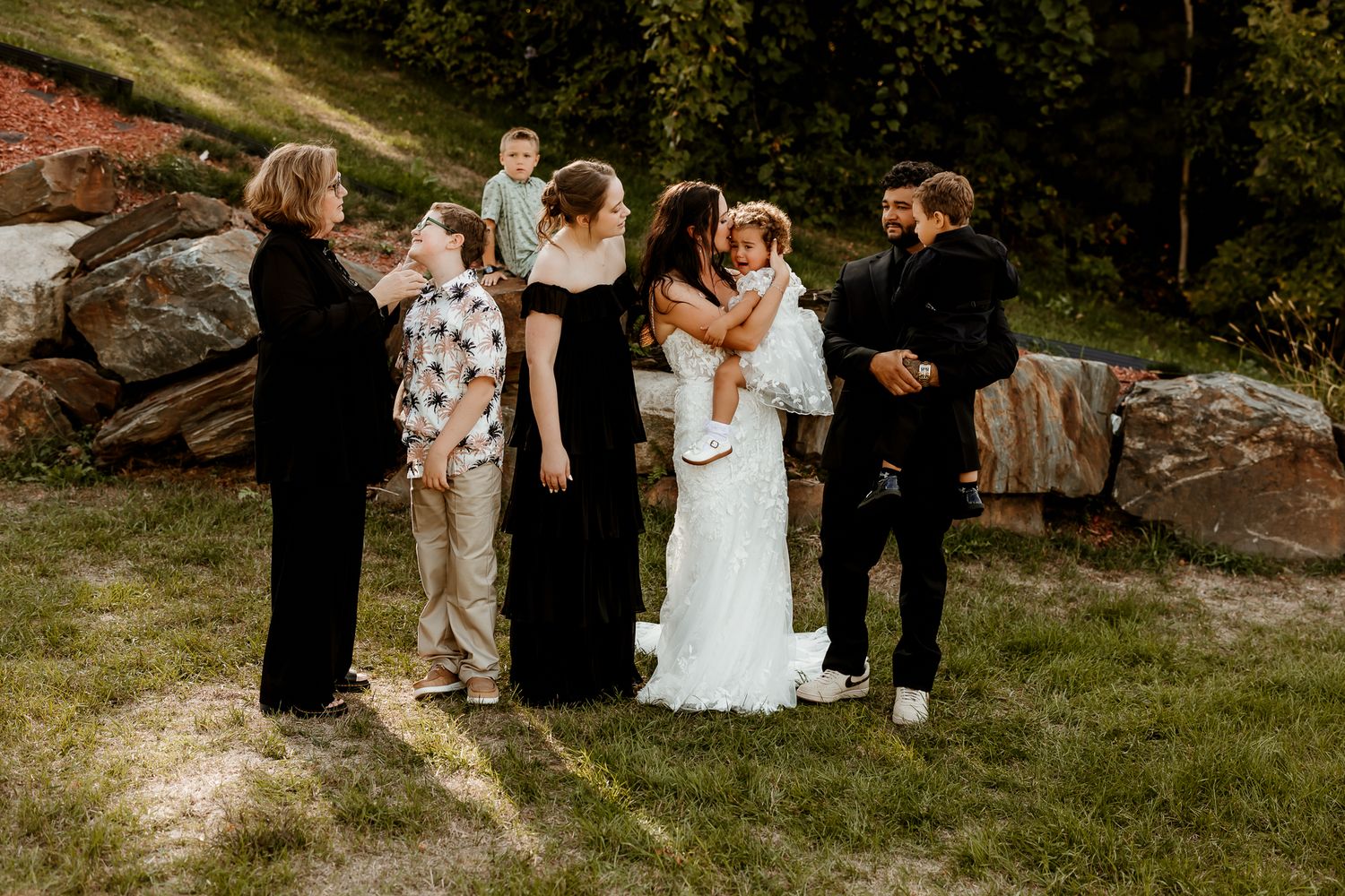 Bride in a white gown embraces a child while family members in dark formal wear gather around her on a grassy hillside beside large rocks.