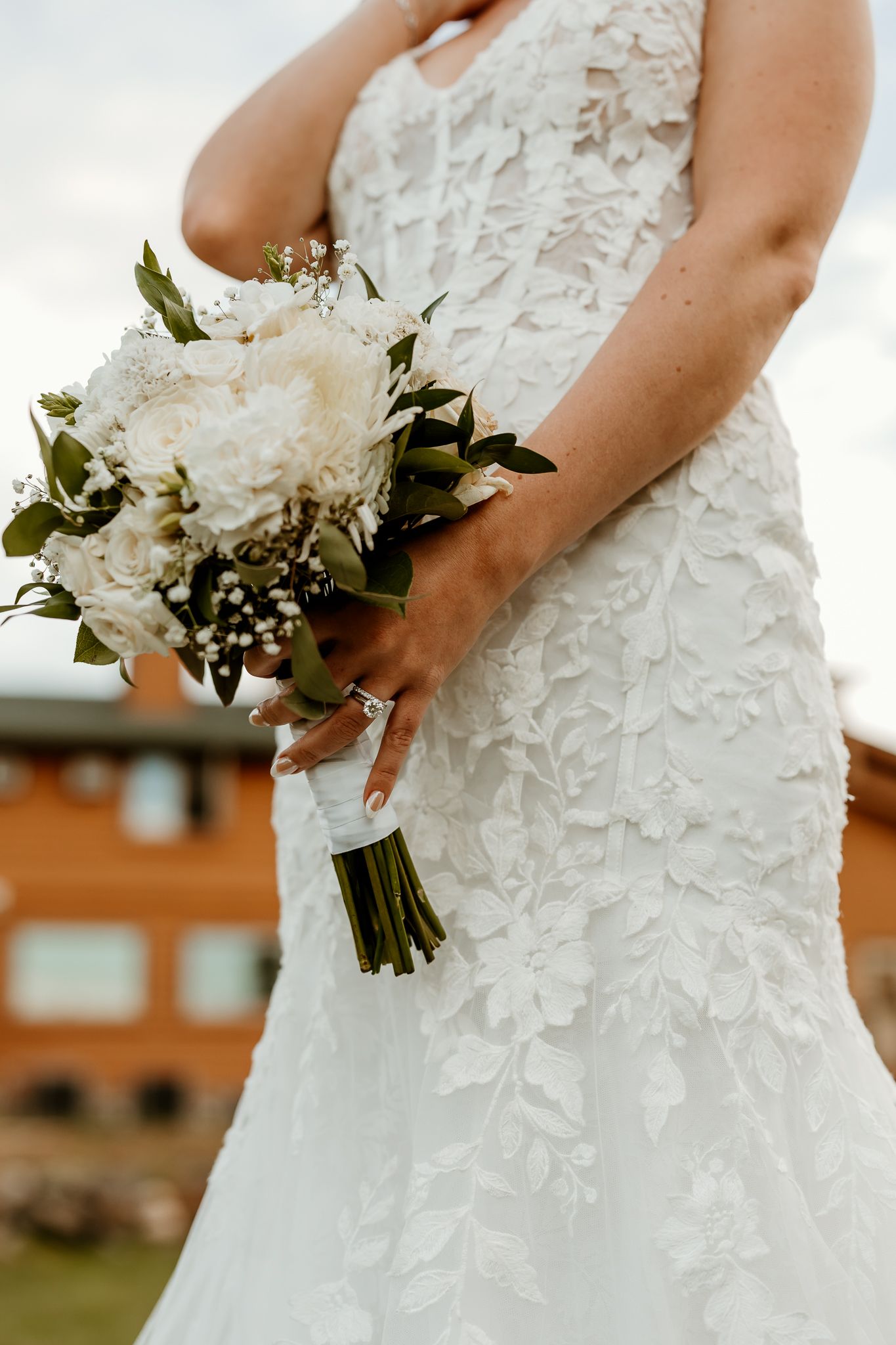 Bride in a white lace wedding gown holding a bouquet of white roses and greenery, with a rustic building in the background.