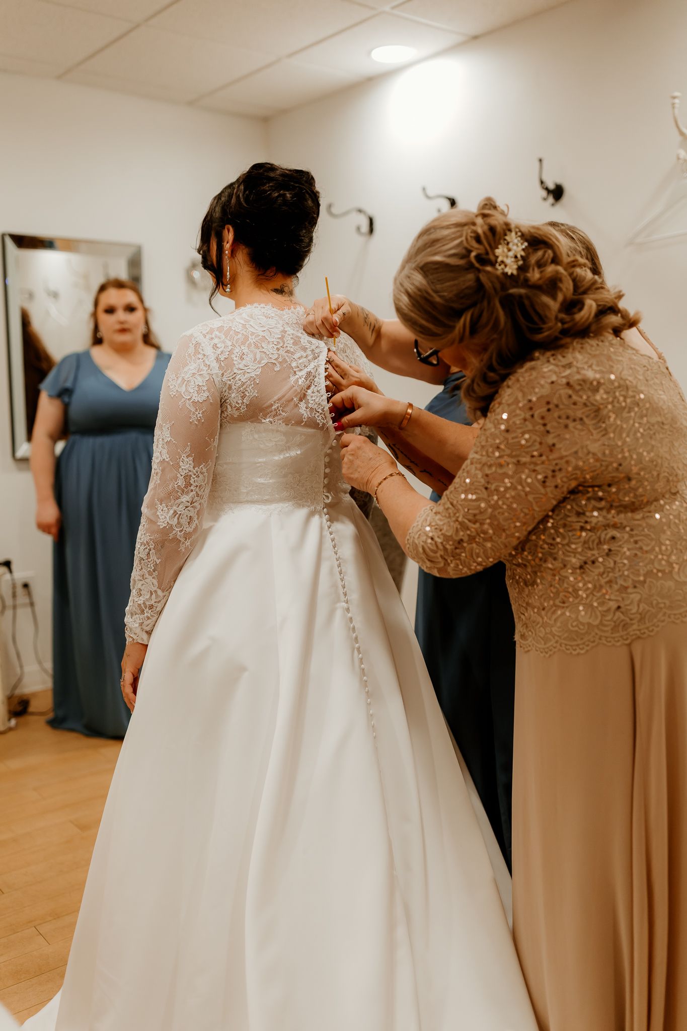 A woman in a sparkly gold dress fastens the buttons on a bride's white lace-sleeved wedding gown while a bridesmaid in blue watches.