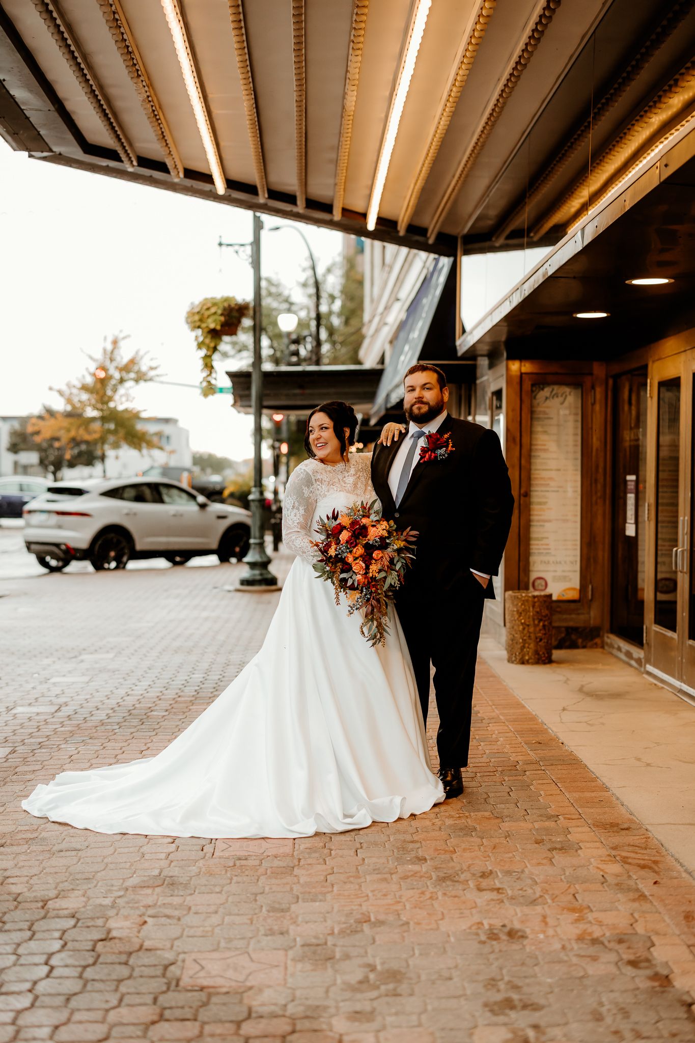 Bride in a long-sleeved lace wedding gown holding an autumn bouquet poses with her groom in a black suit under a theater marquee.