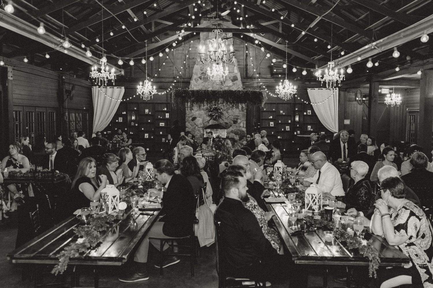Black and white photo of wedding guests seated at long reception tables beneath chandeliers in a rustic barn venue.