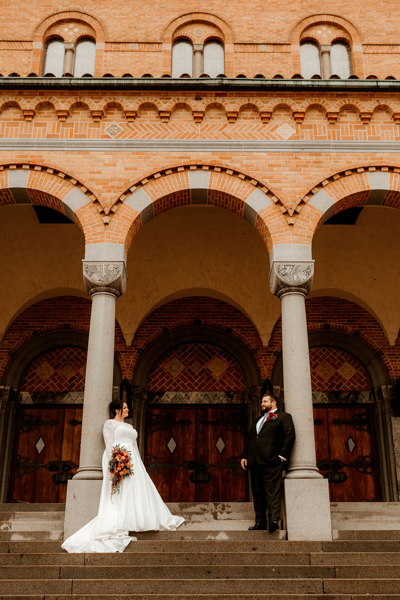 Bride in a white gown holding a bouquet and groom in a dark suit stand on stone steps before a red brick building with arched columns.