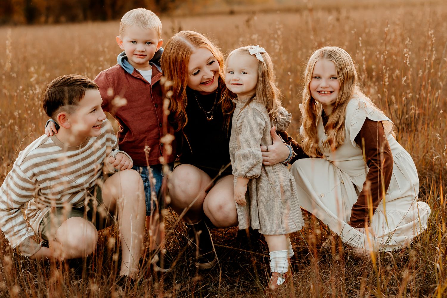 A smiling red-haired woman poses with four happy children in a golden grassy field during a warm sunset family photo session.