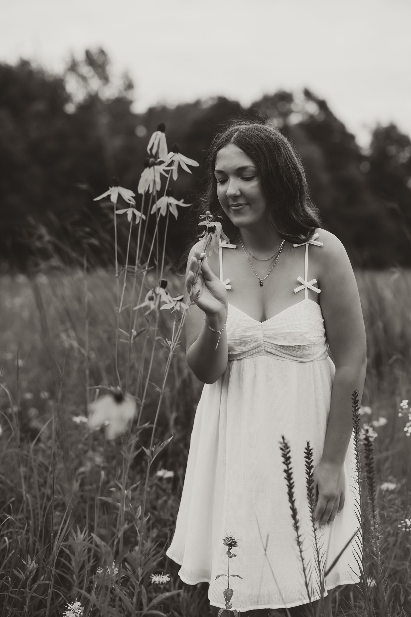 Black and white photo of a young woman in a white sundress smiling while holding wildflowers in a meadow.