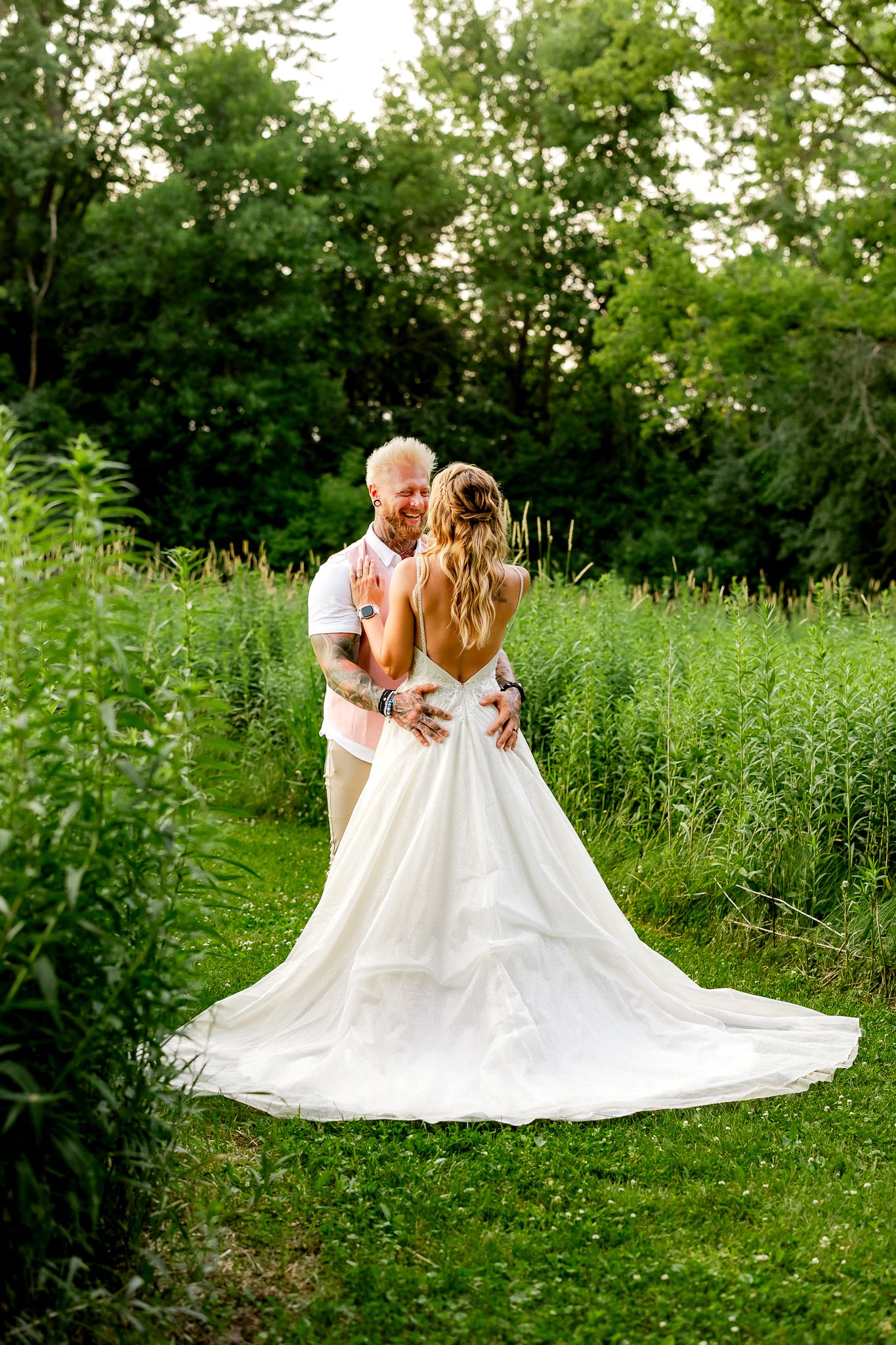Bride and groom embrace in lush green garden, her white gown train spread elegantly on the grass.