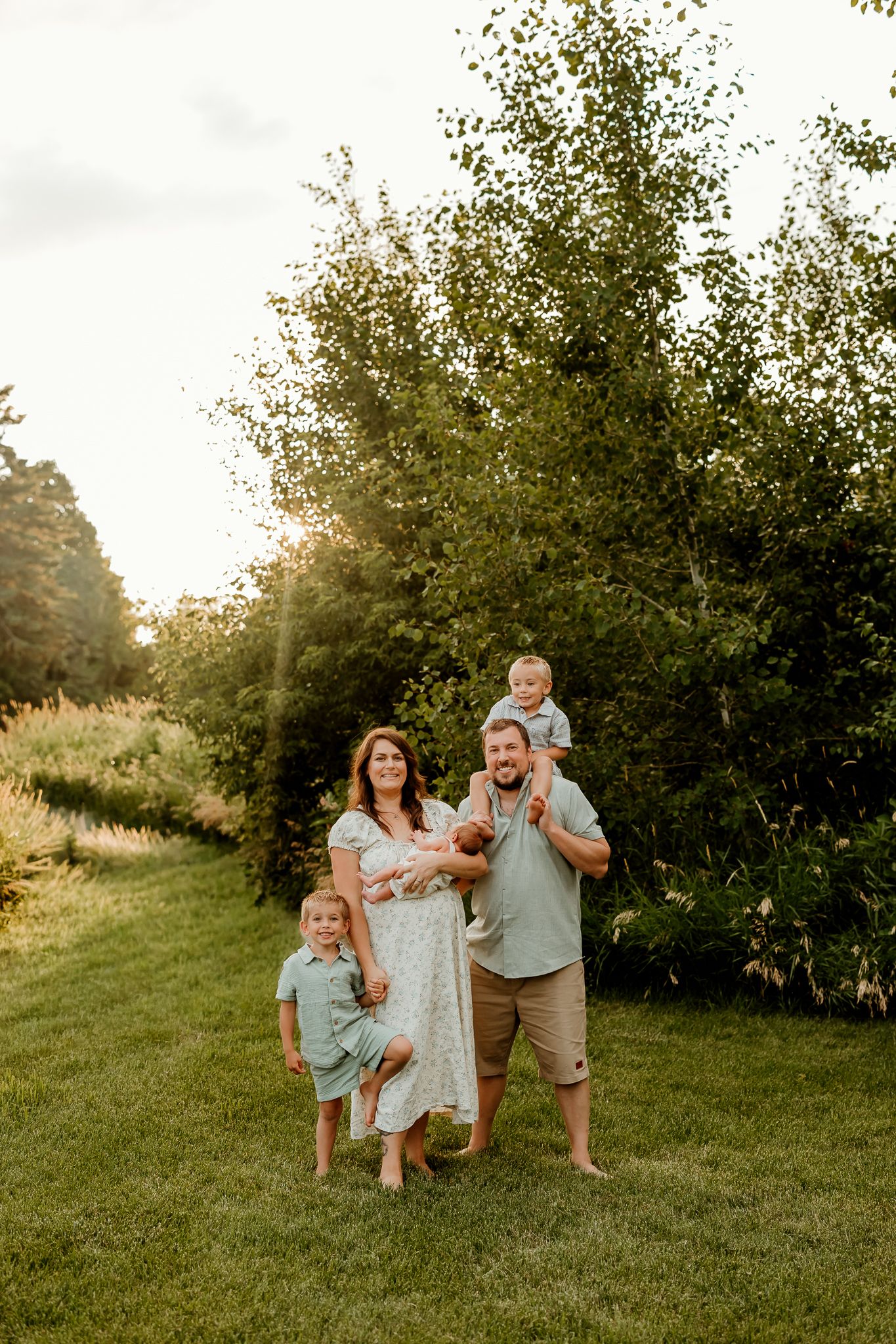 Family of four poses outdoors at golden hour, parents holding babies amid lush green trees on a sunlit lawn.