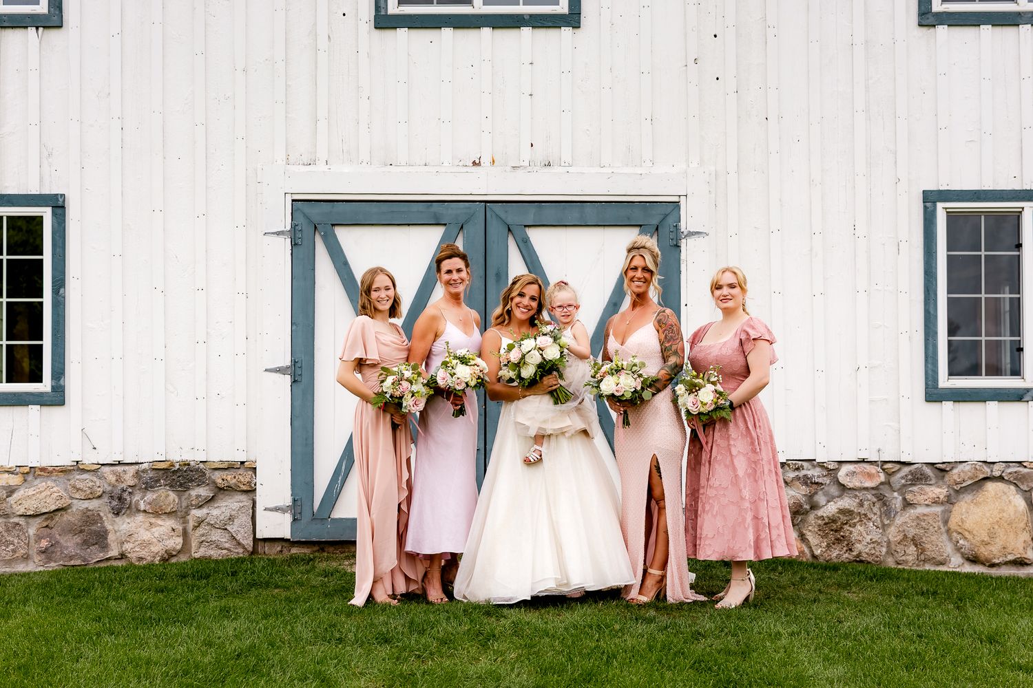 Bride in white gown with four bridesmaids in pink dresses holding bouquets outside a white barn wedding venue.