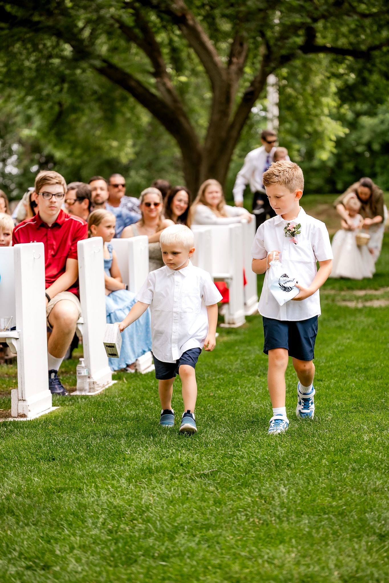 Two young boys in white shirts walk down an outdoor wedding aisle on green grass, with seated guests watching.