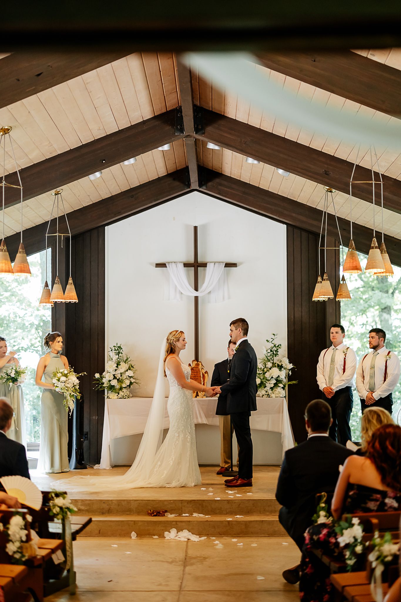 Bride and groom exchange vows at elegant chapel altar with wooden cross, flanked by wedding party and floral arrangements.