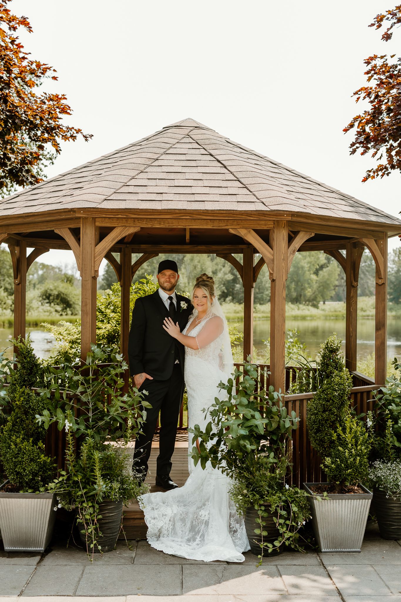 Bride and groom posing in front of a wooden gazebo surrounded by lush greenery at an outdoor wedding venue.