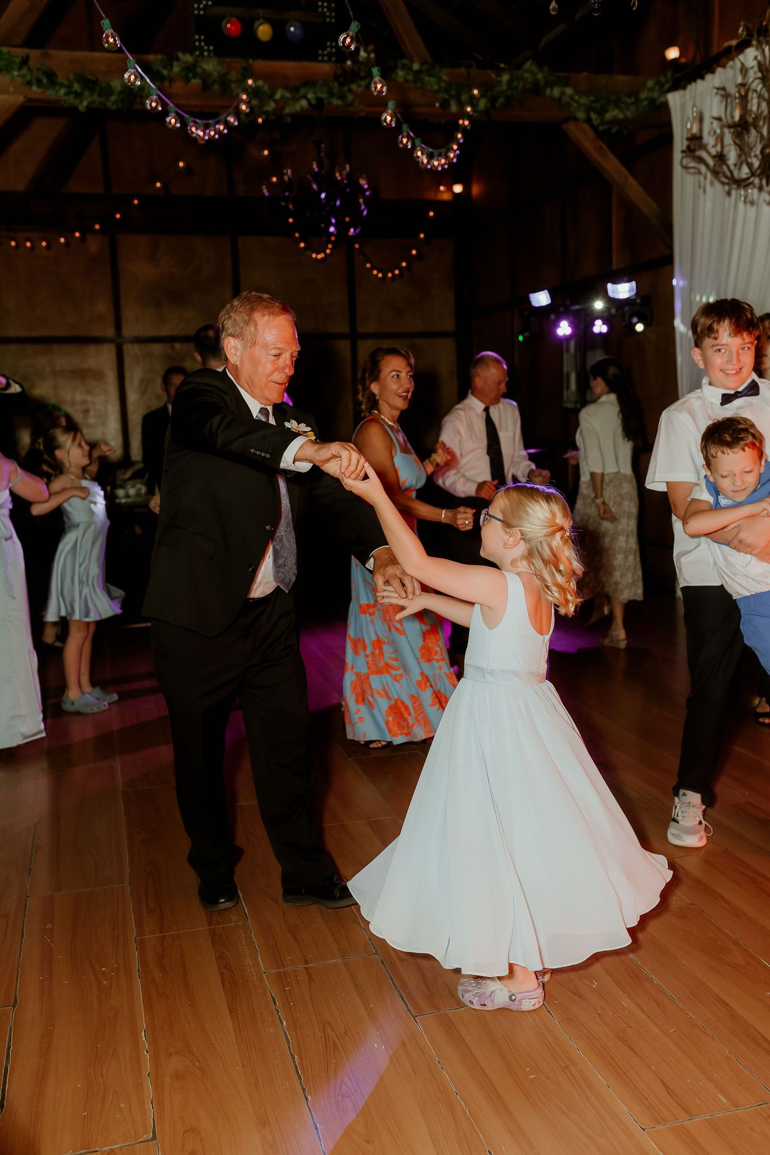 Man in suit dancing with young girl in white dress at wedding reception on wooden dance floor.