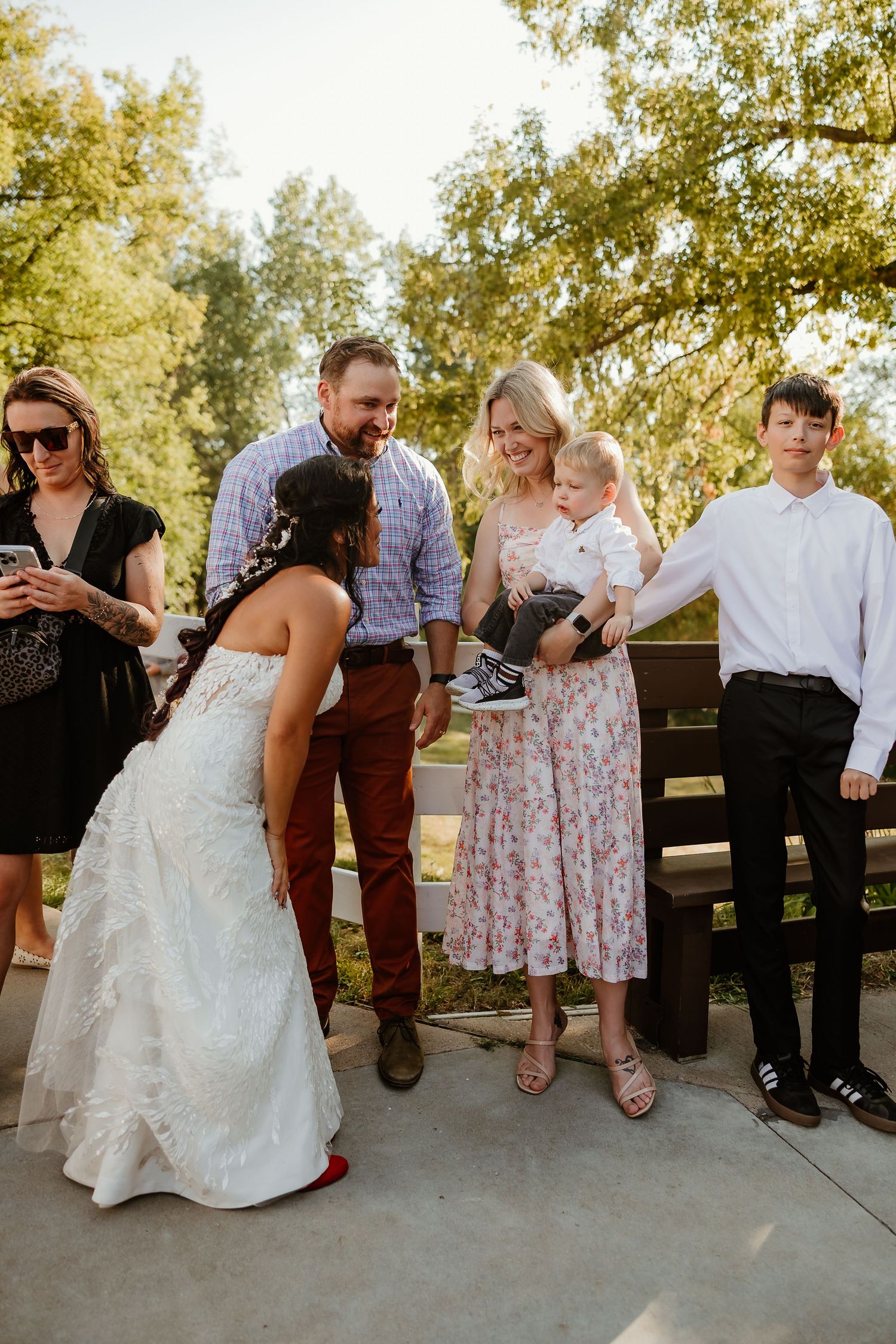 Bride in white gown leans to greet a baby held by a woman in floral dress at an outdoor wedding gathering.