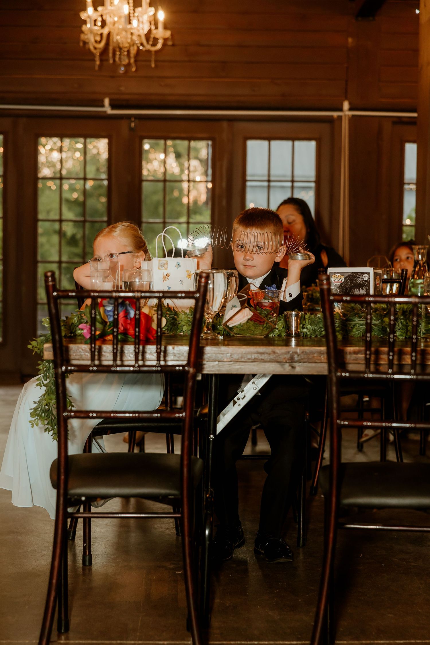 Two children sit at a decorated wedding reception table inside a rustic wood-paneled venue with chandelier lighting.
