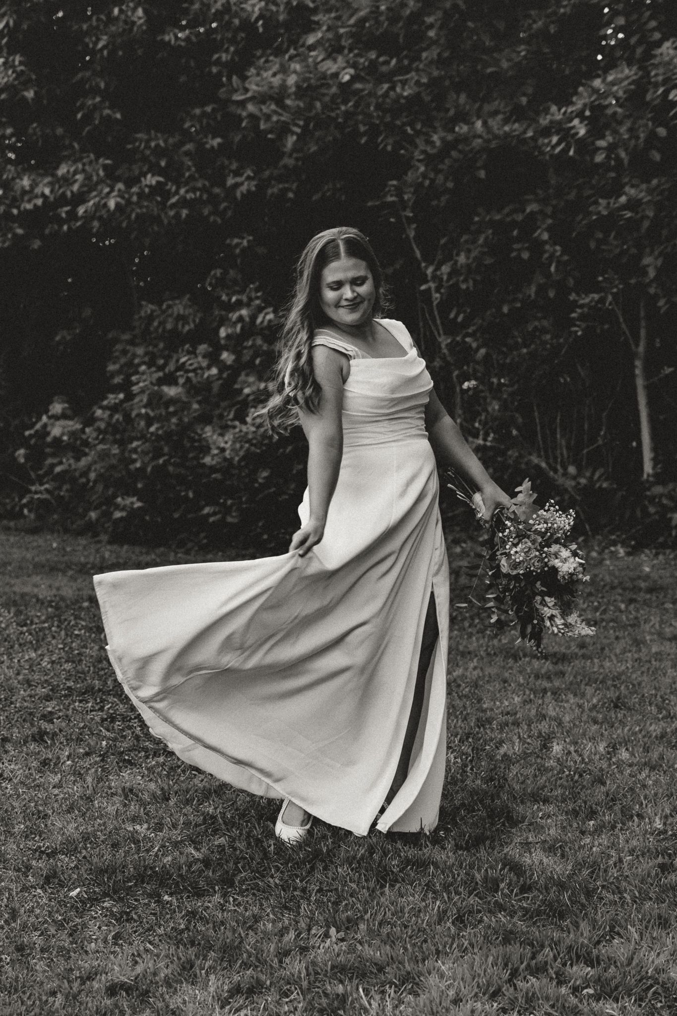 Black and white photo of a smiling bride in a flowing gown holding a bouquet outdoors.
