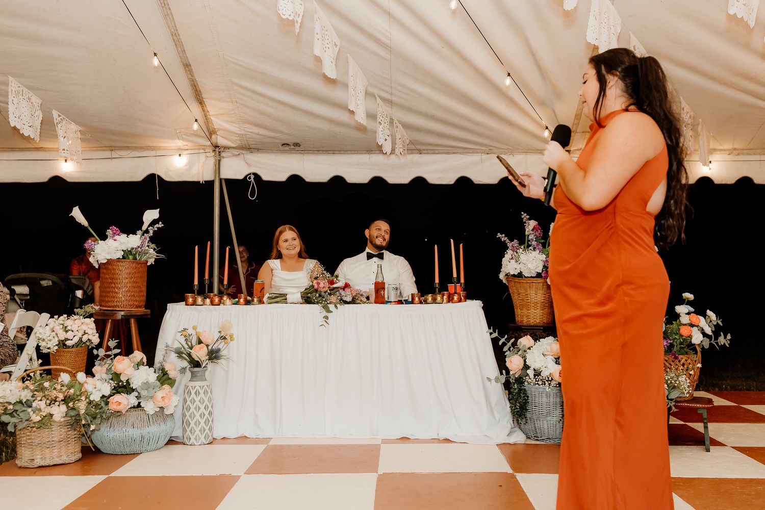 Woman in orange dress giving a speech at a wedding reception under a tent with the bride and groom seated at the head table.
