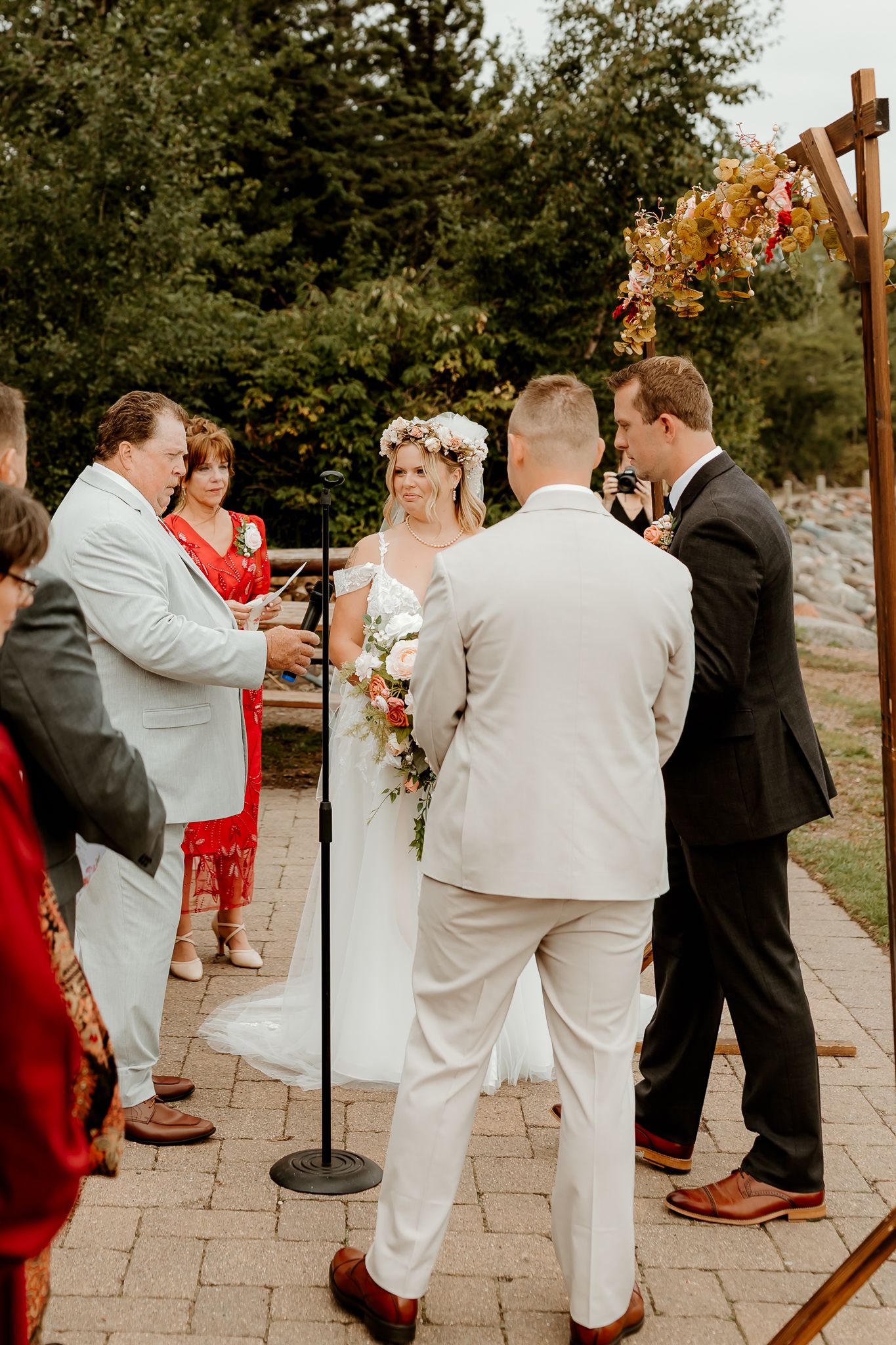Outdoor wedding ceremony with bride in floral crown, groom in white suit, officiant, and guests on brick patio.