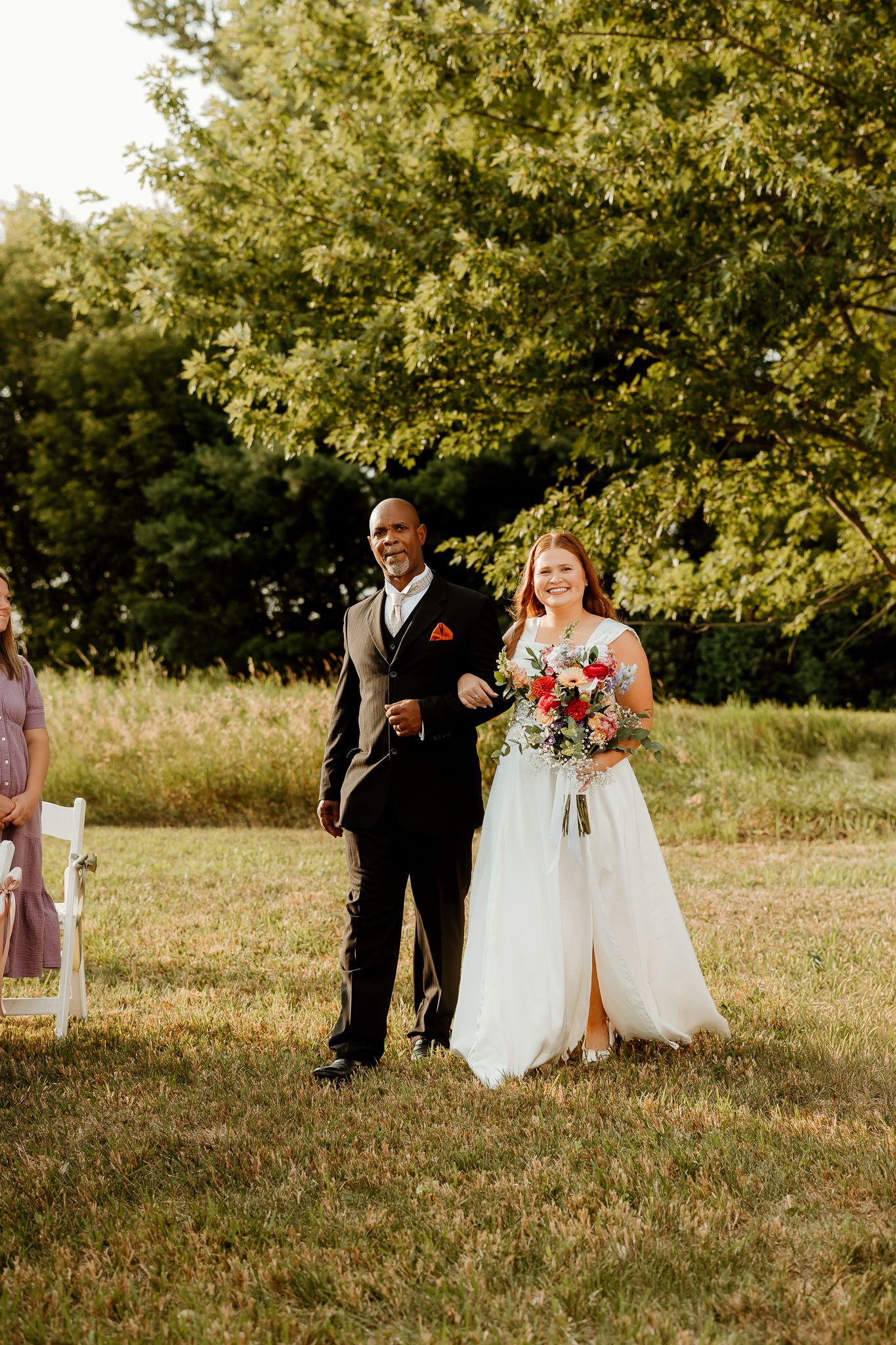 Bride in white gown holding colorful bouquet walks down outdoor aisle with escort at sunny garden wedding ceremony.