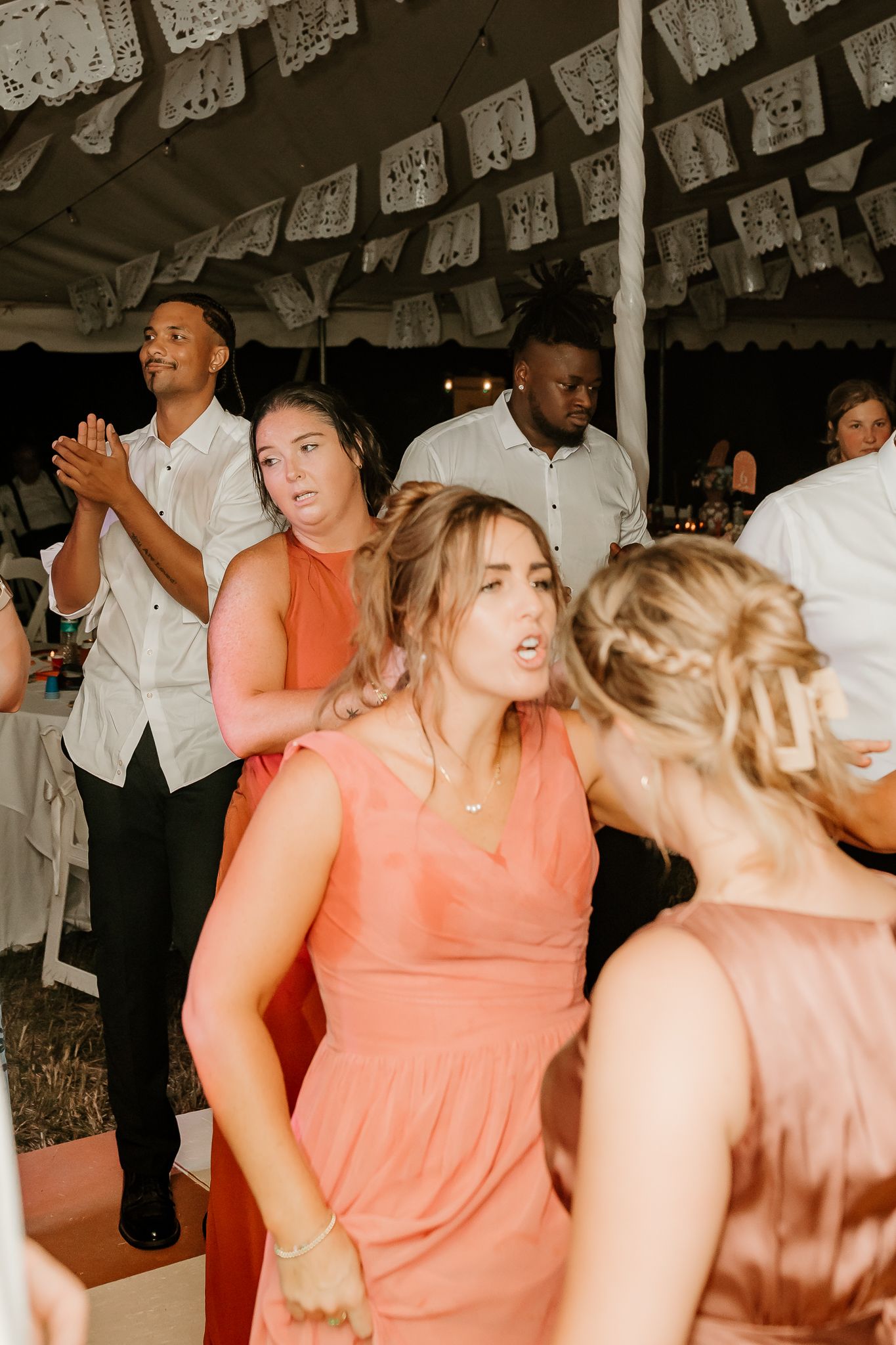 Bridesmaids in coral dresses dancing at an outdoor wedding reception under a decorated tent.
