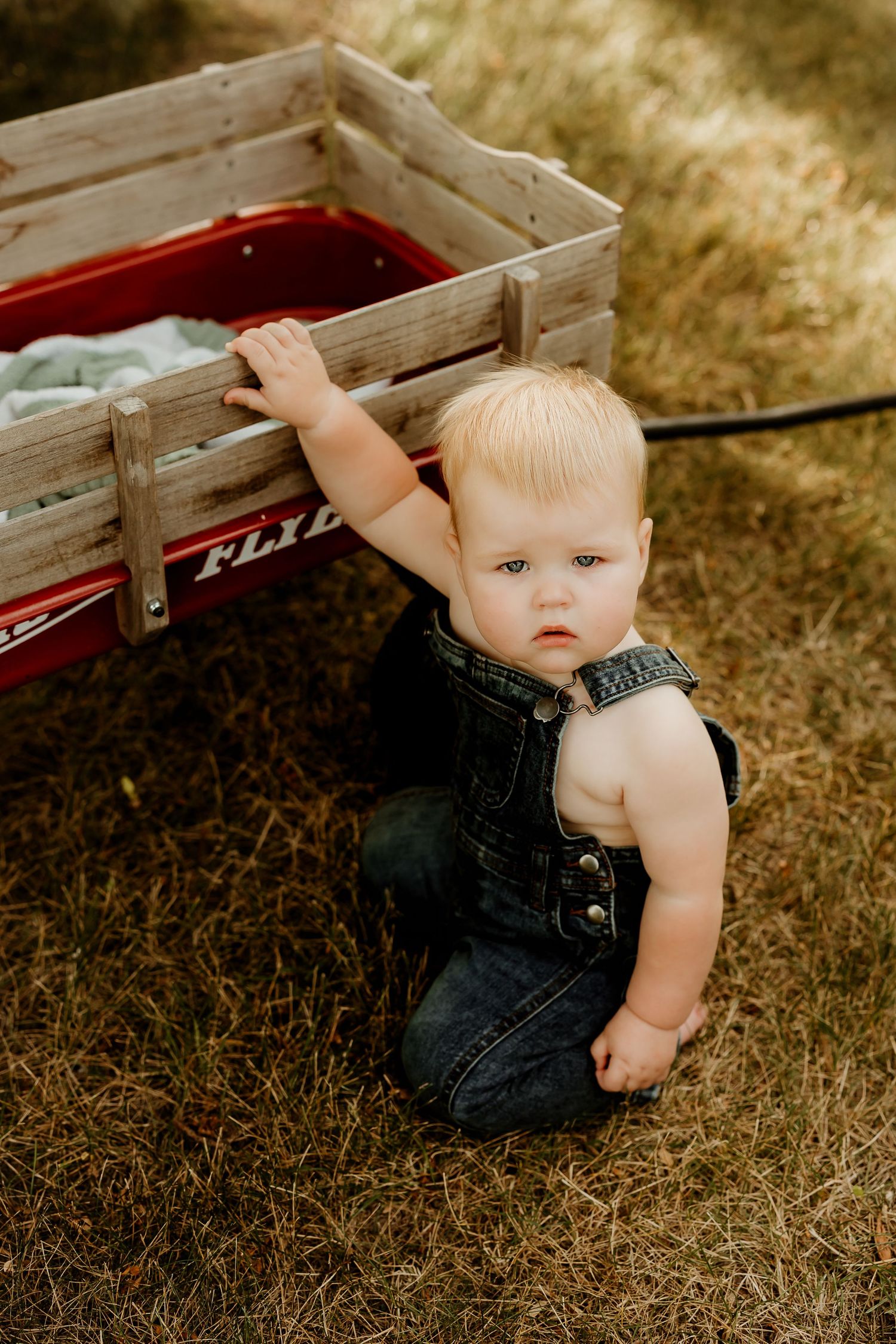 Blonde toddler in denim overalls sits on grass beside a red Radio Flyer wagon outdoors.