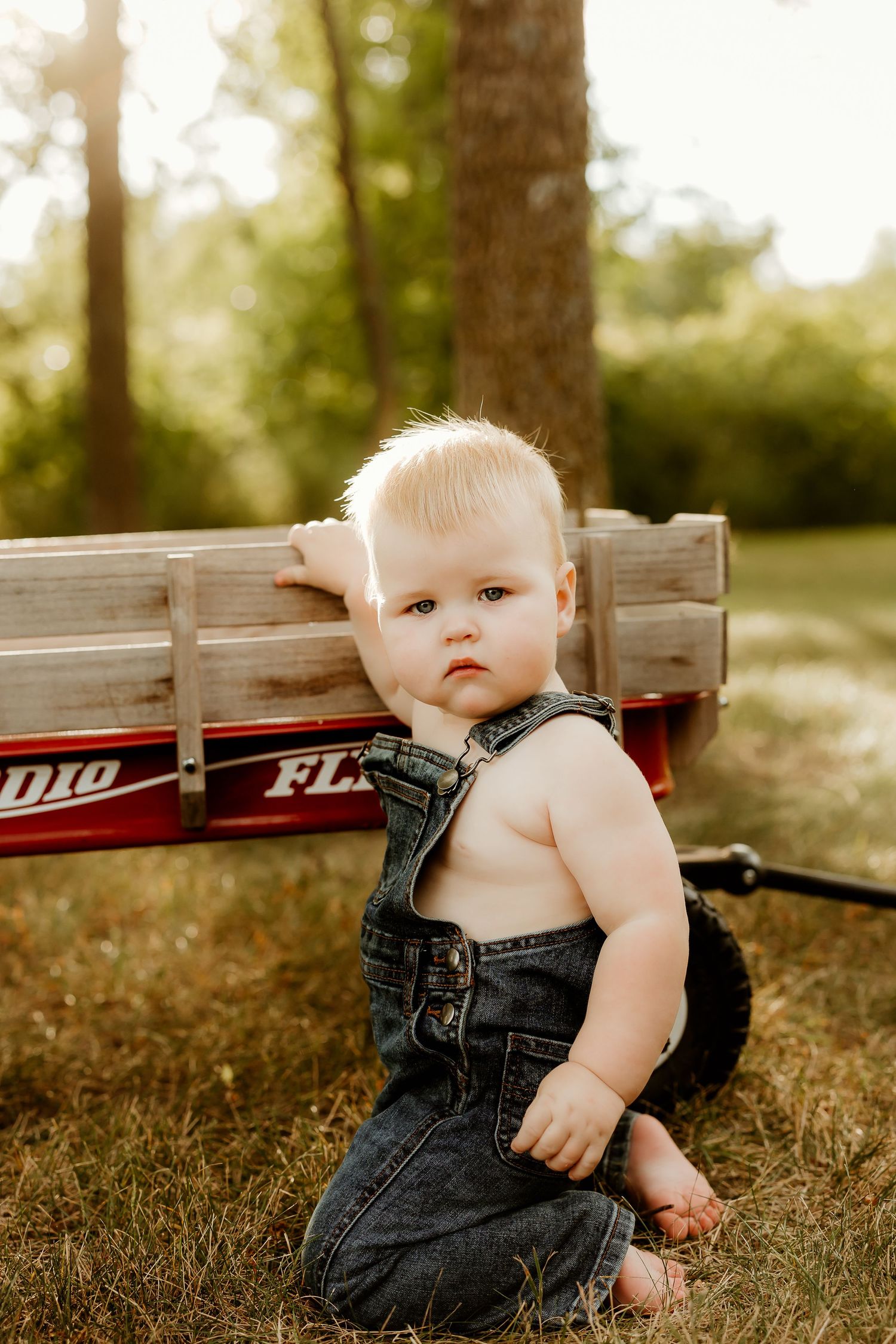 Blonde toddler in denim overalls sitting on grass beside a red Radio Flyer wagon outdoors.